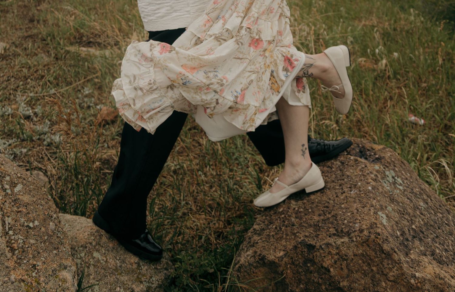Couple stepping on rocks, wearing floral dress and black boots with beige flats, outdoor nature setting.