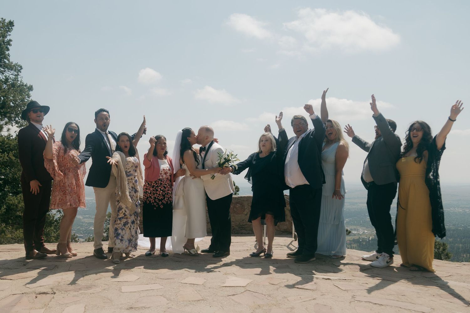 Wedding group photo outdoors on scenic overlook with guests raising hands in celebration under blue sky.