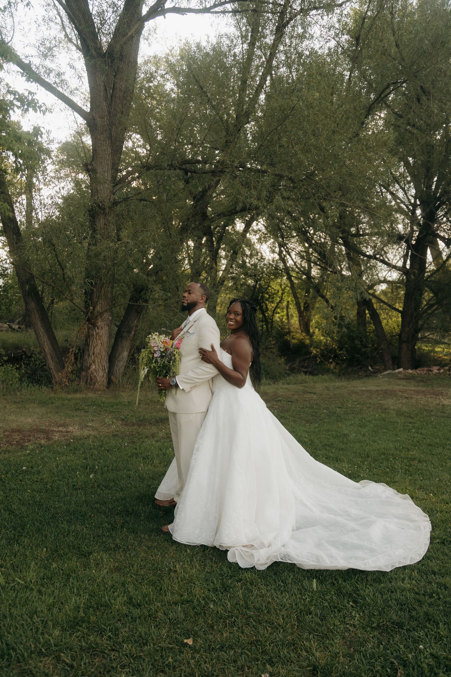 Black bride and groom pose together in a lush green outdoor setting, bride holding a bouquet in a flowing white gown.