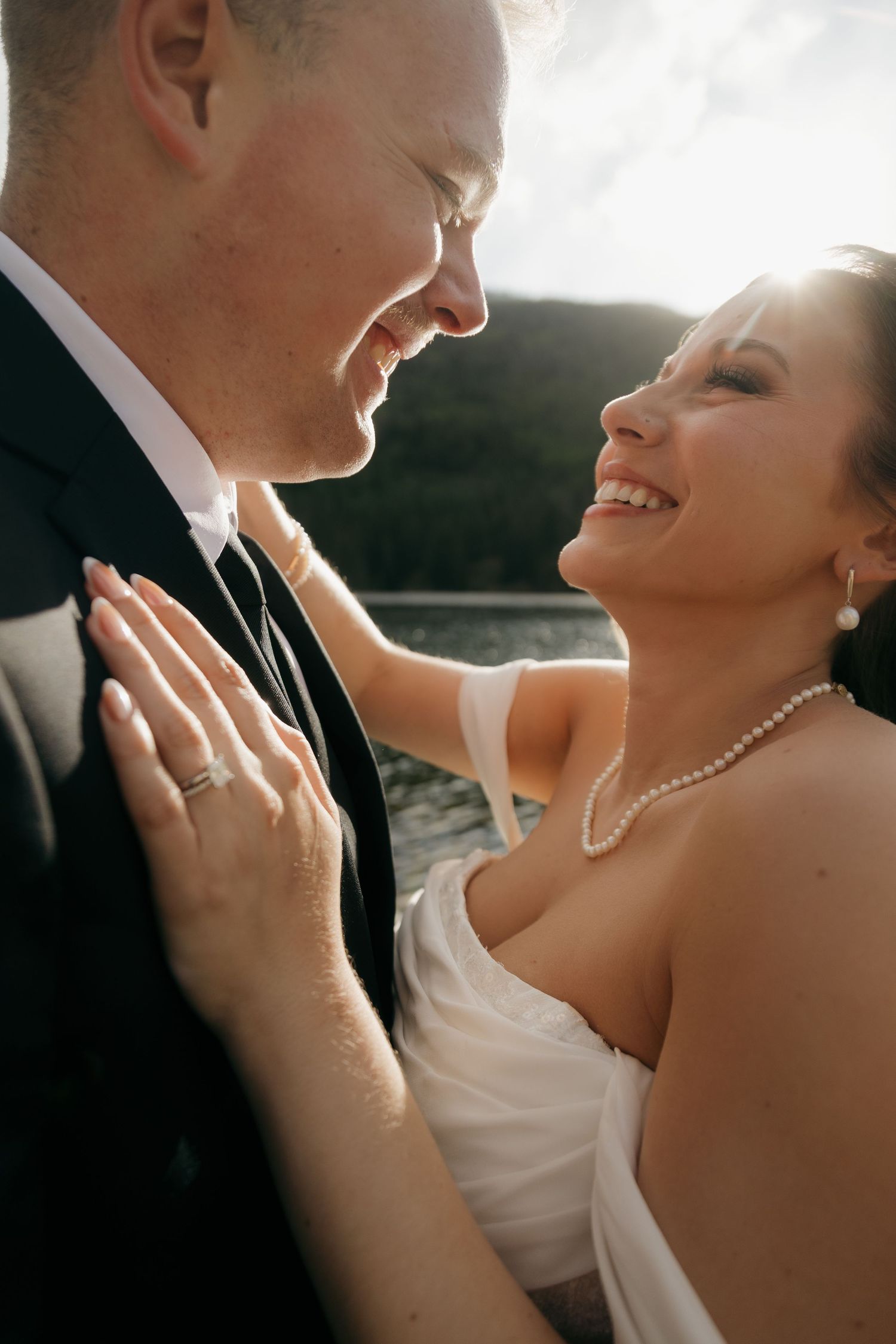 Smiling bride in white dress and pearl necklace gazes at groom in black suit, showcasing wedding ring outdoors.