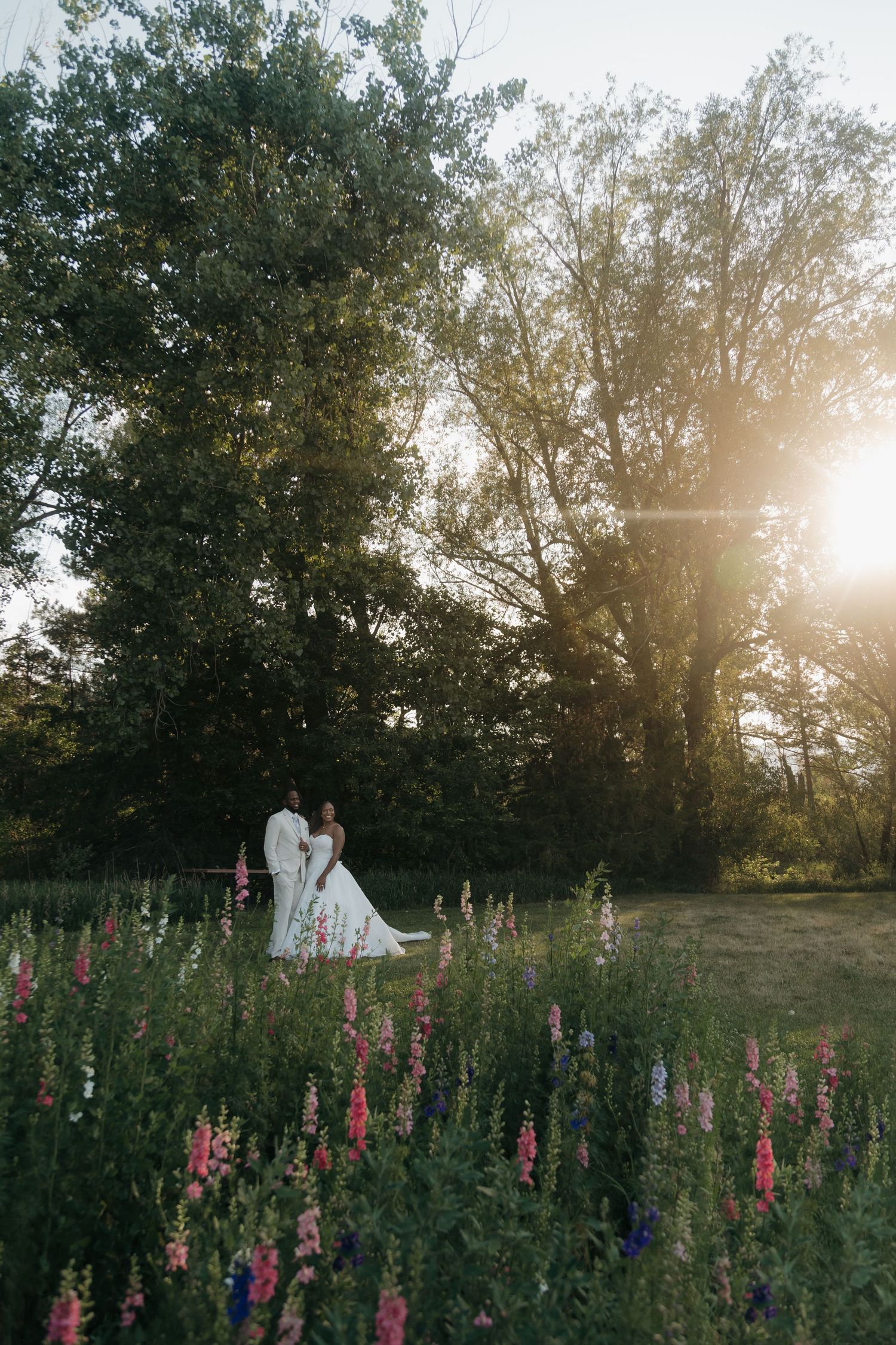 Bride and groom embrace in a sunlit garden with colorful wildflowers and tall trees at golden hour.