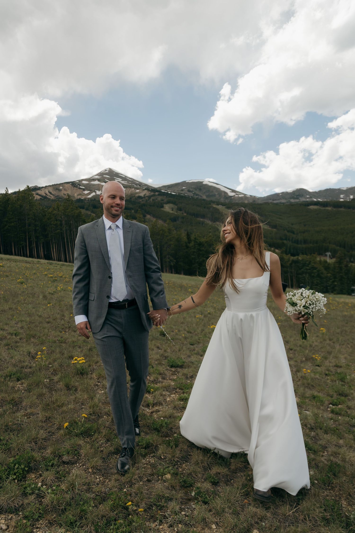 Bride and groom walking hand in hand through mountain meadow, bride in white gown holding white bouquet.
