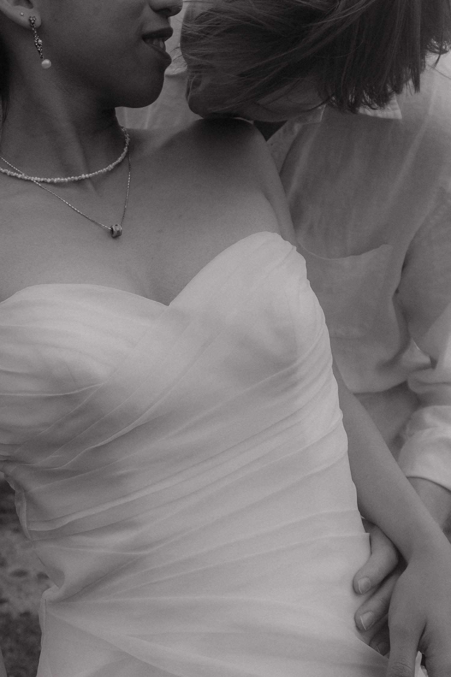 Black and white close-up of bride in strapless sweetheart gown with delicate necklace, embraced by her partner.