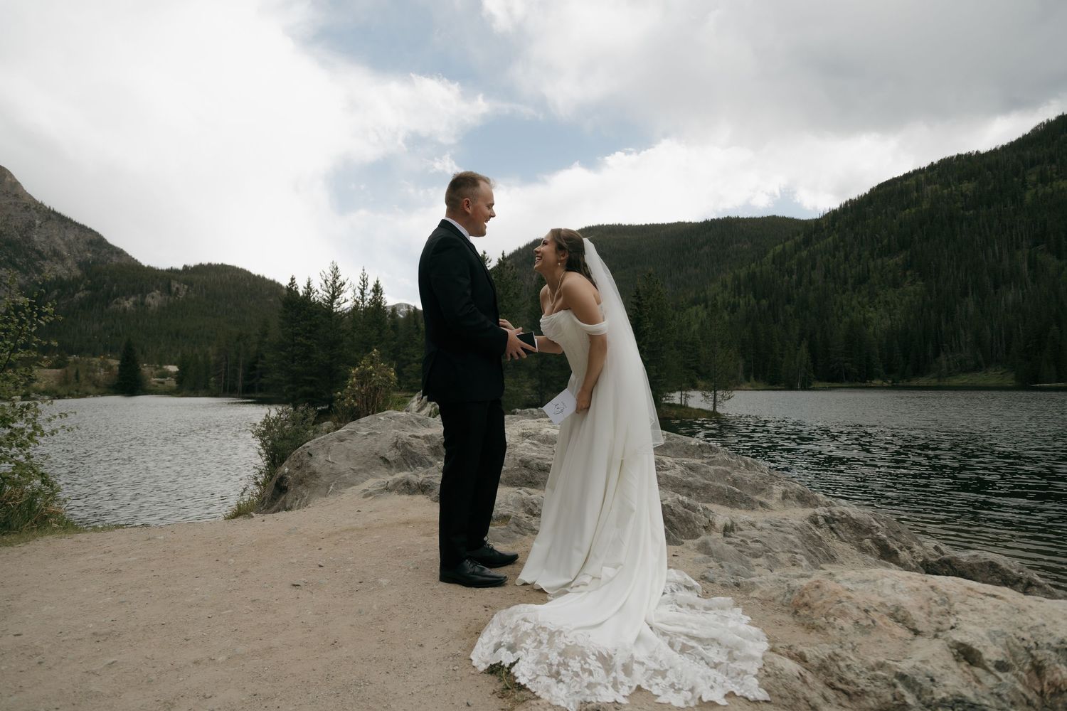 Bride and groom exchanging vows lakeside with mountain forest backdrop on their outdoor wedding day.