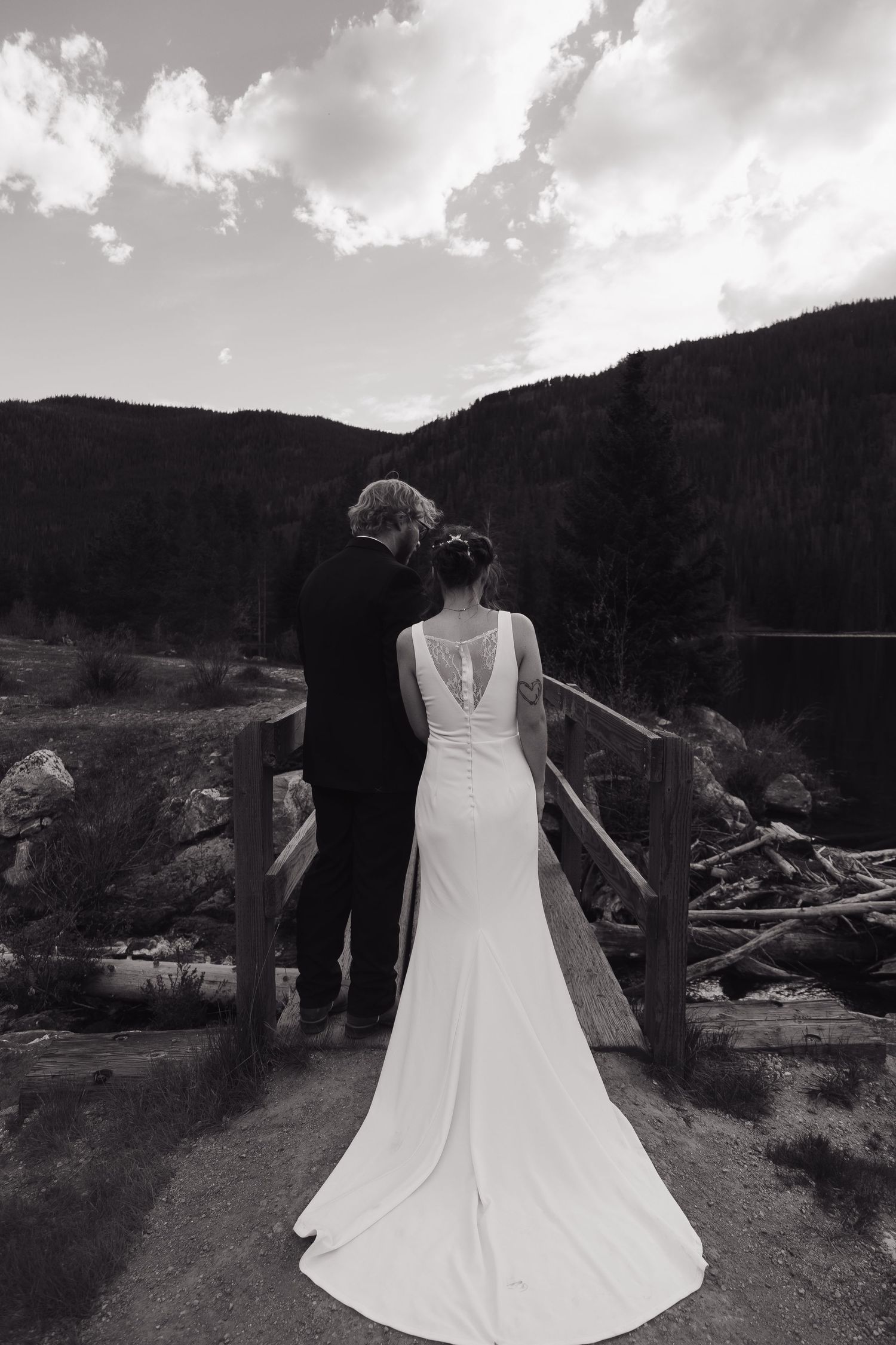 Black and white photo of bride and groom standing on wooden bridge with mountain forest backdrop.