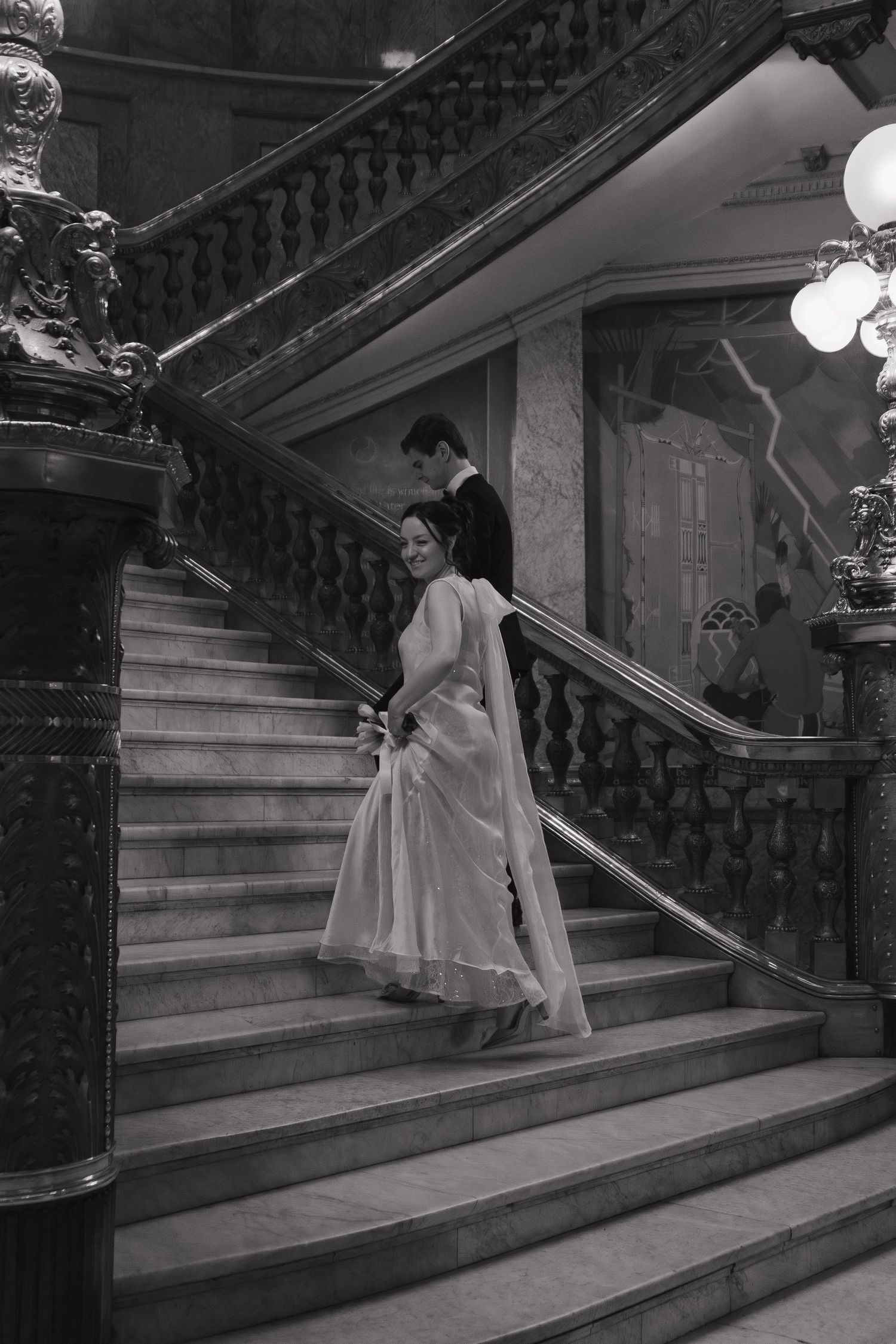 A couple descends an elegant grand staircase with ornate railings in a vintage black and white photograph.