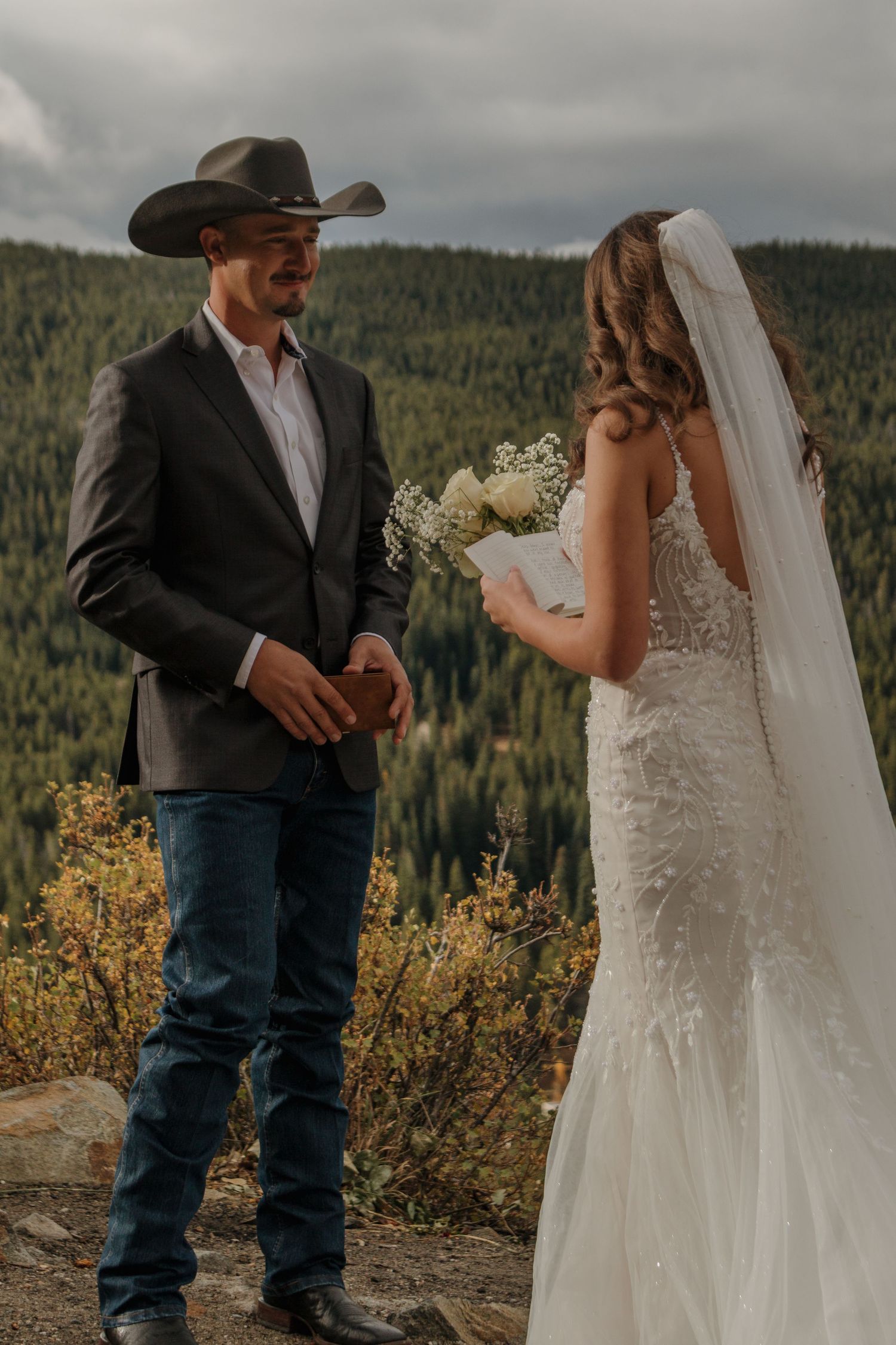 A couple stands together against a mountain vista with golden wildflowers in the foreground.