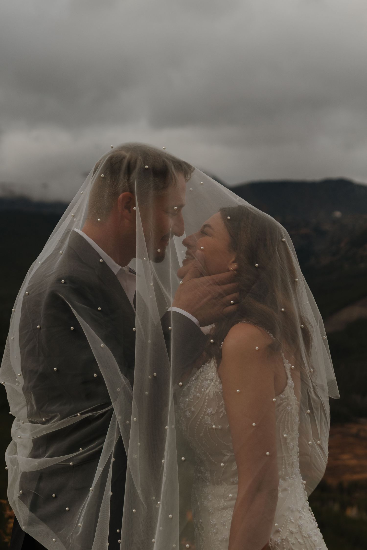 A romantic wedding moment captured under a veil against a moody mountain backdrop.