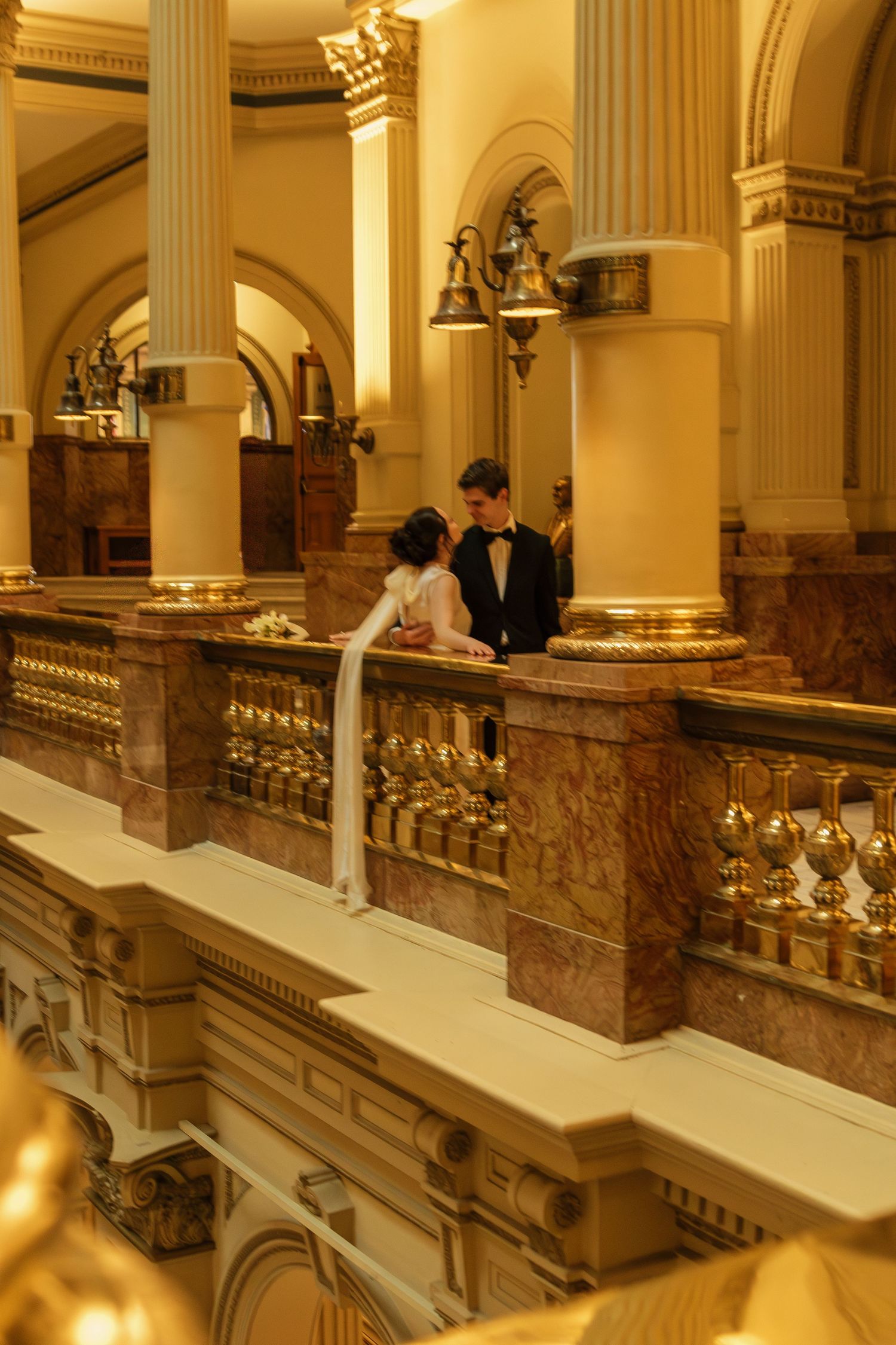 A couple stands in an ornate classical building interior with golden columns and marble details.