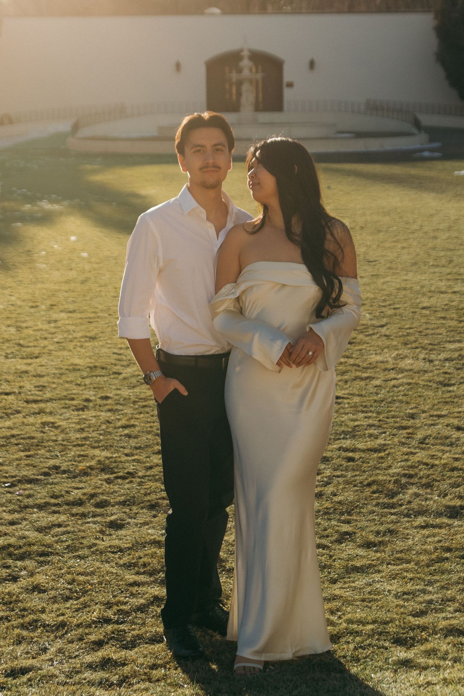 A romantic sunset portrait on a grassy lawn with a white building and mountains in the distance.