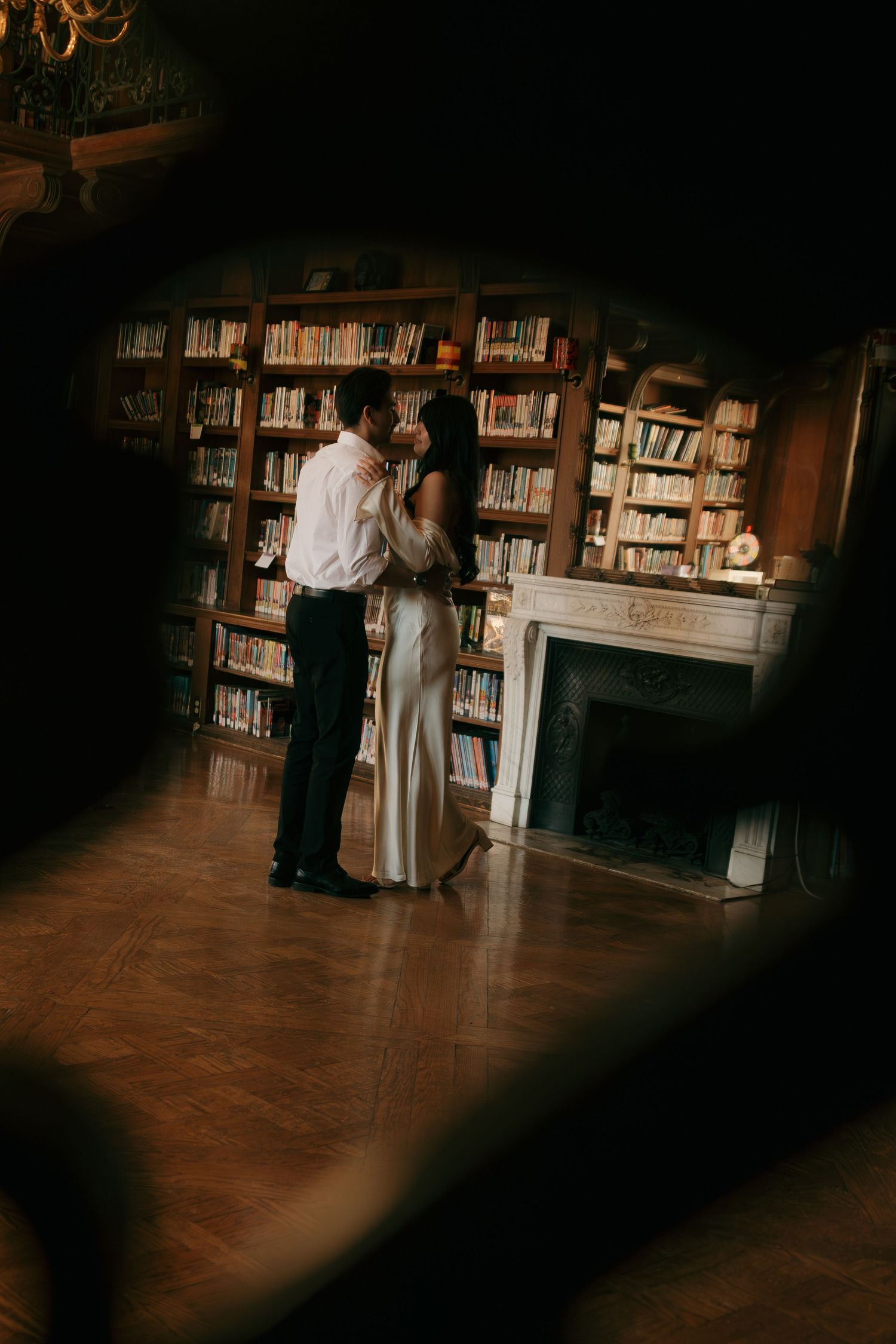 A romantic couple dances intimately in a dimly lit library surrounded by bookshelves and wooden floors.