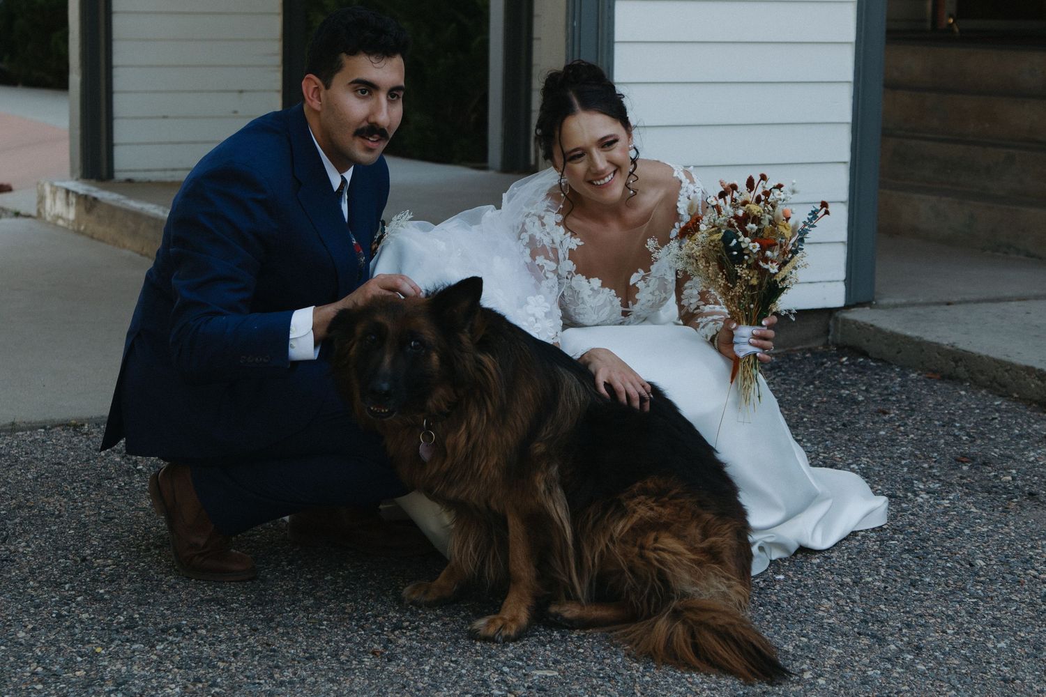 Wedding couple posing with their dog on a gravel path outside a white house.