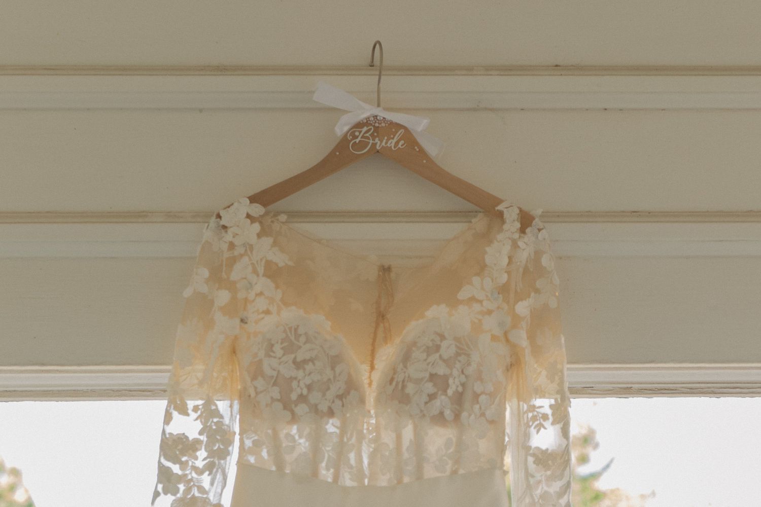 Delicate lace wedding dress hanging on a wooden hanger against a light beige wall near white window trim.