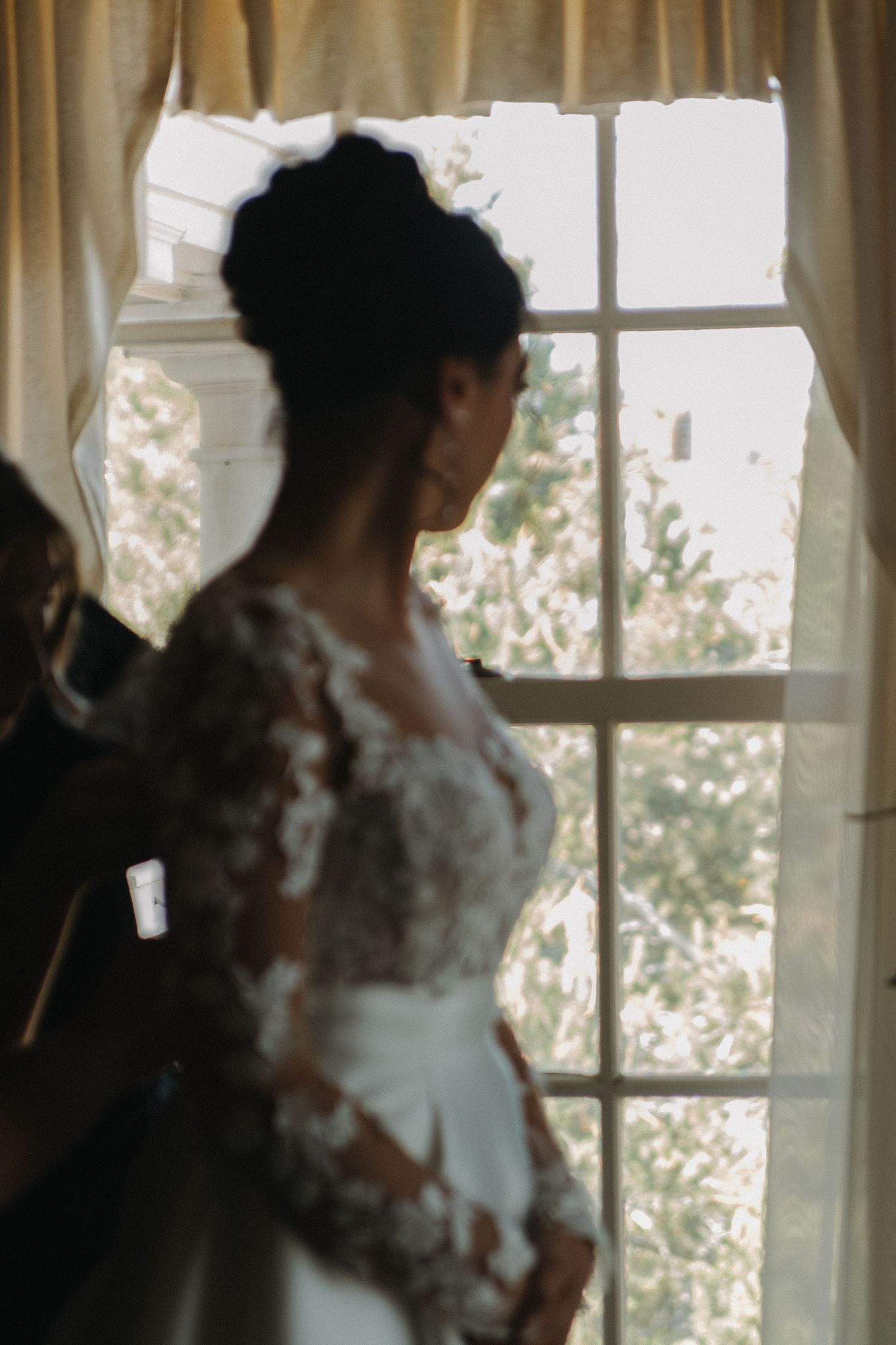 A silhouetted figure in a wedding dress stands by a sunlit window with sheer curtains.