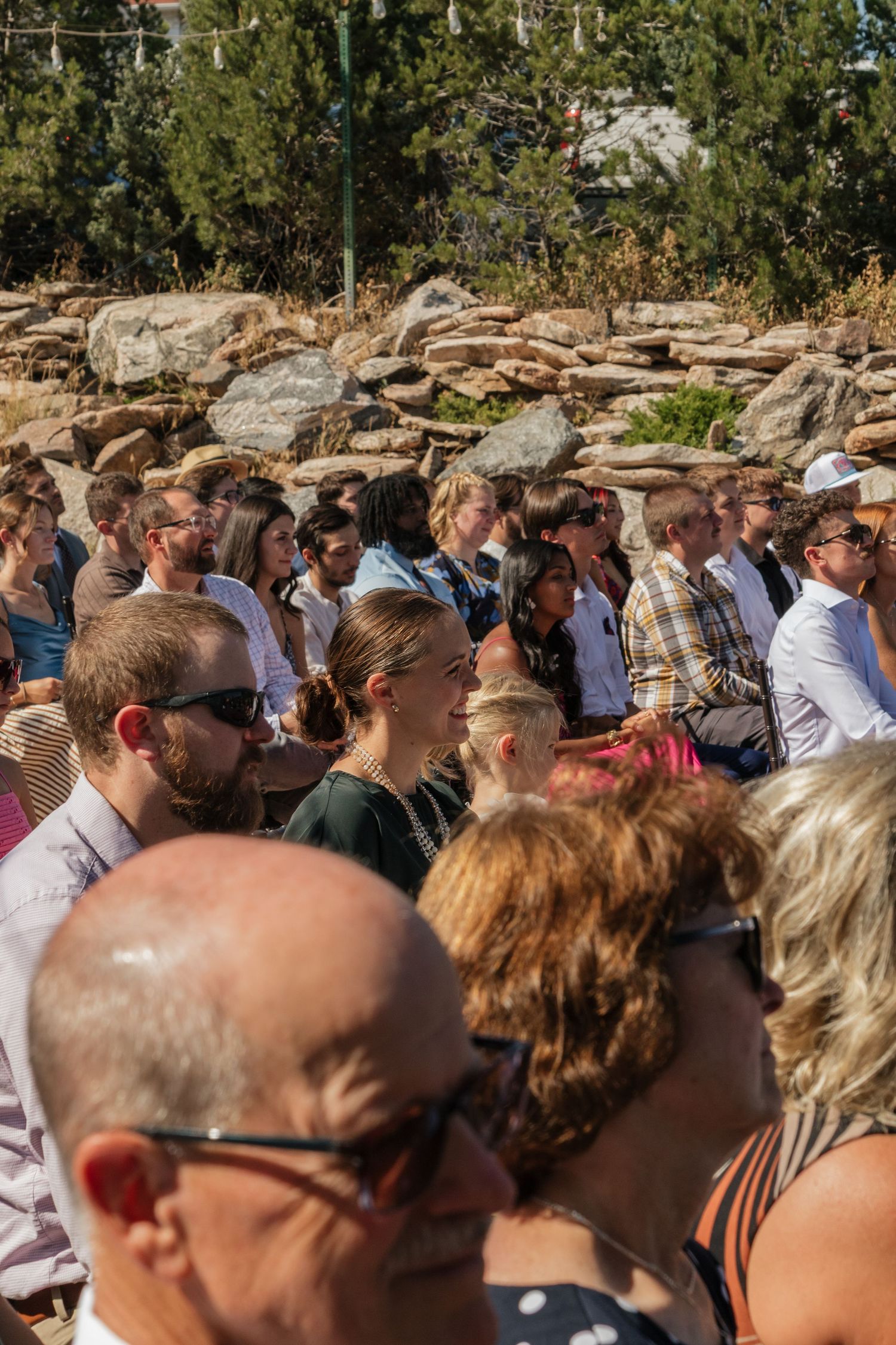Large crowd of wedding guests gathered outdoors with rocky landscape and trees in the background.