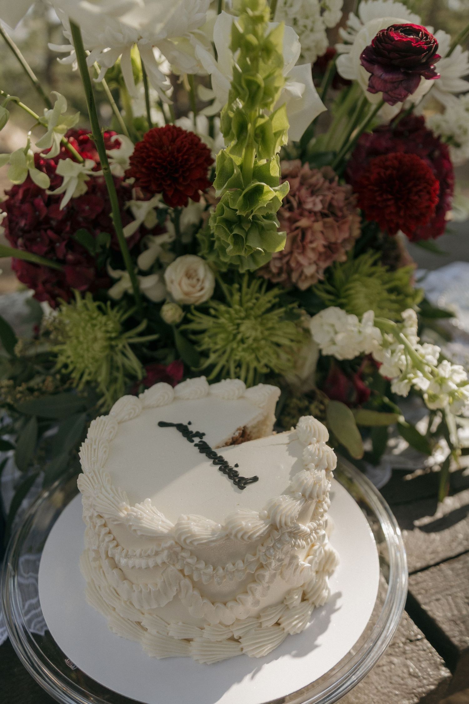 Elegant white wedding cake with piped frosting displayed in front of a lush floral arrangement.