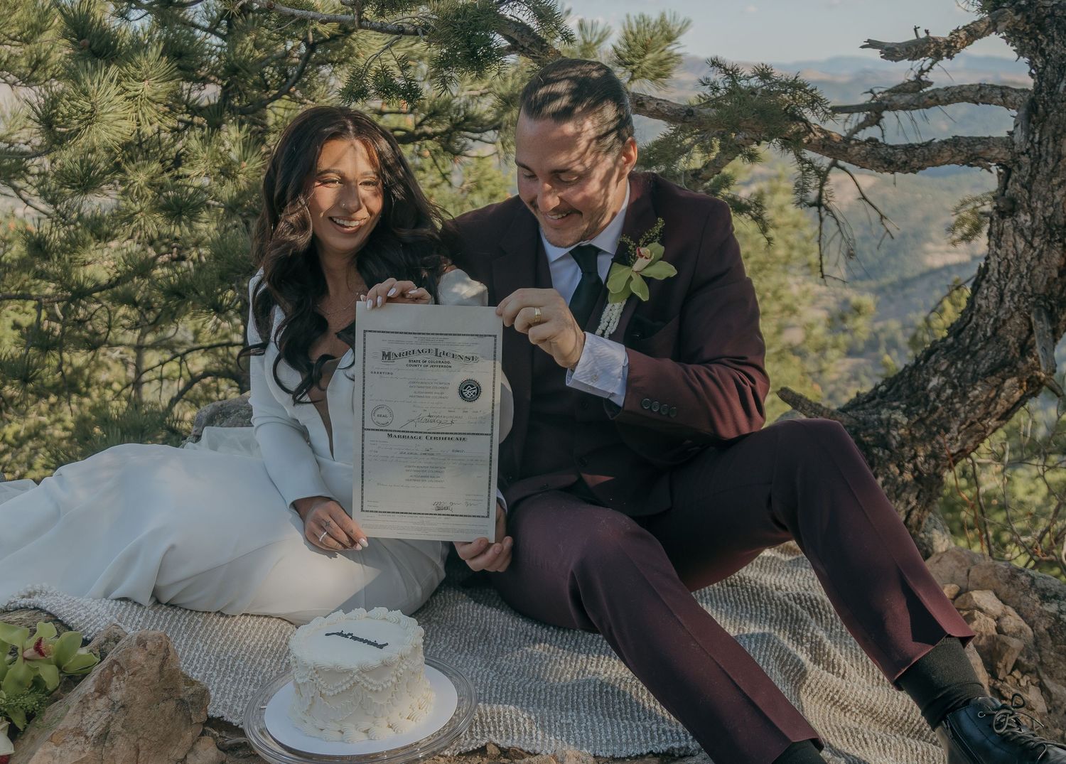 Bride and groom sit outdoors holding their marriage certificate beside a small white wedding cake under pine trees.