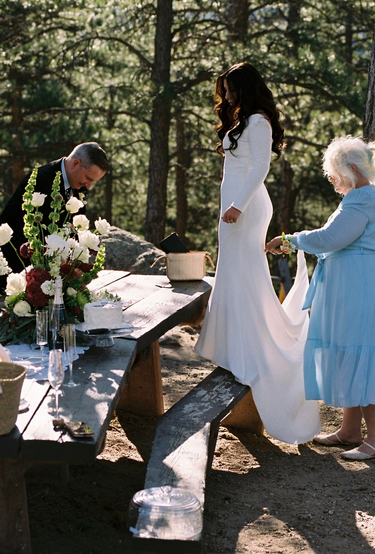 Bride in elegant white gown stands at a rustic outdoor table with floral arrangements during reception.