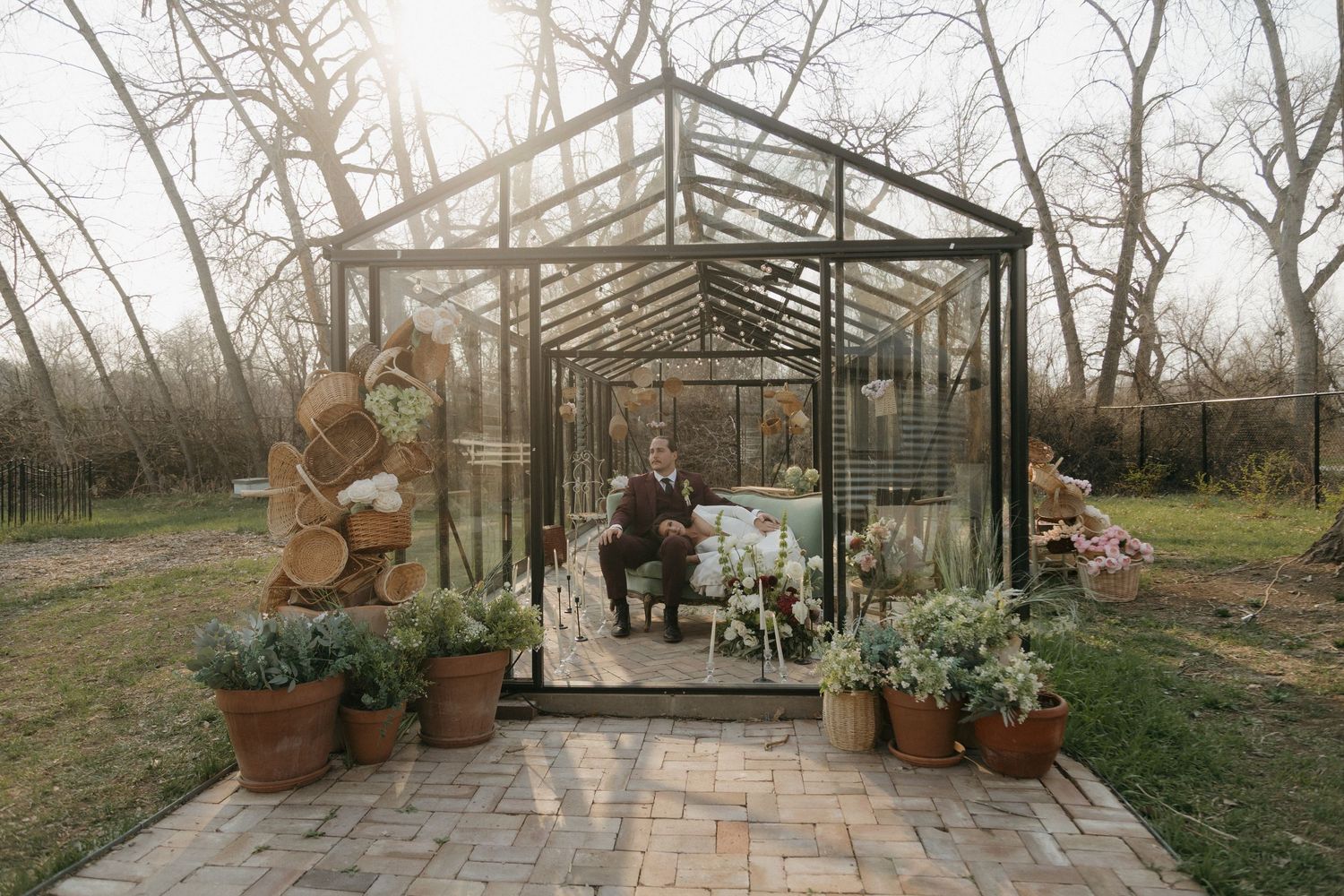 Groom sits inside a glass greenhouse wedding venue surrounded by potted plants and rustic wood decor.