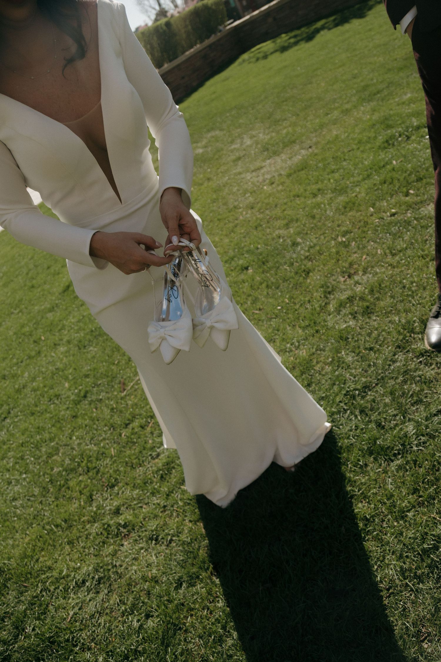 Bride in white long-sleeve wedding dress holding white bow heels while standing on green grass outdoors.