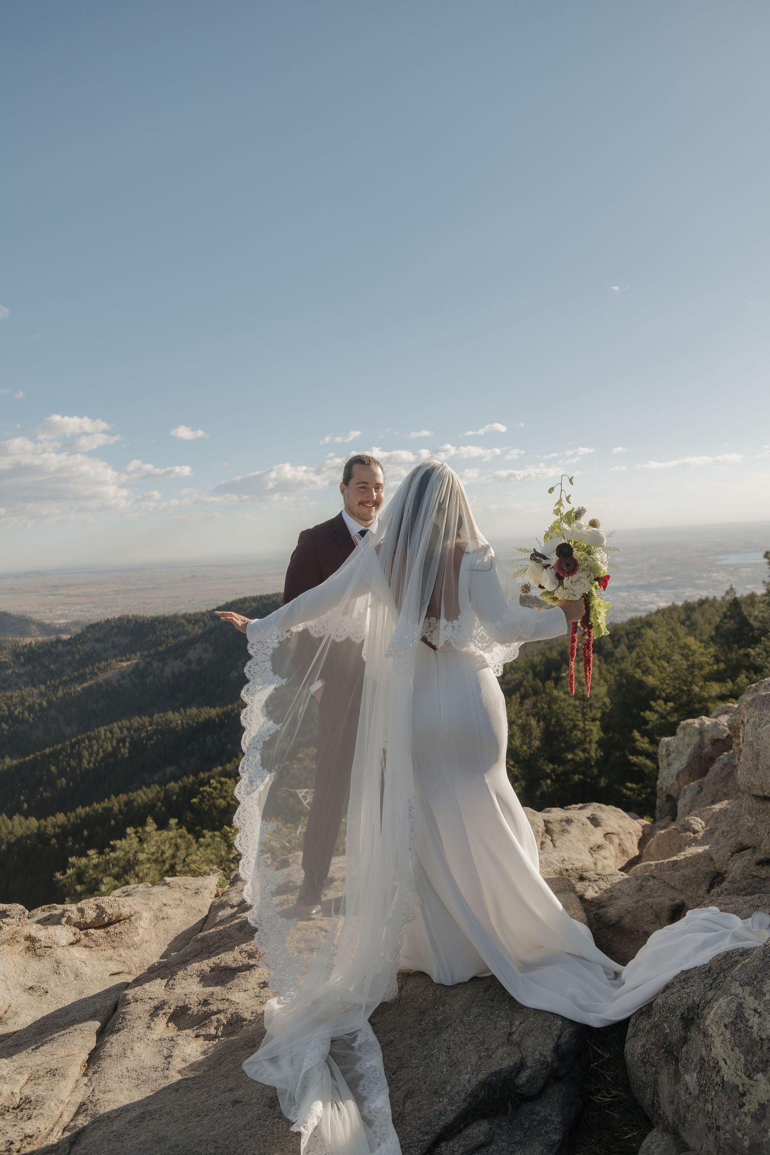 Bride in a flowing white gown and veil holds a bouquet while facing groom in burgundy suit on a mountain summit.