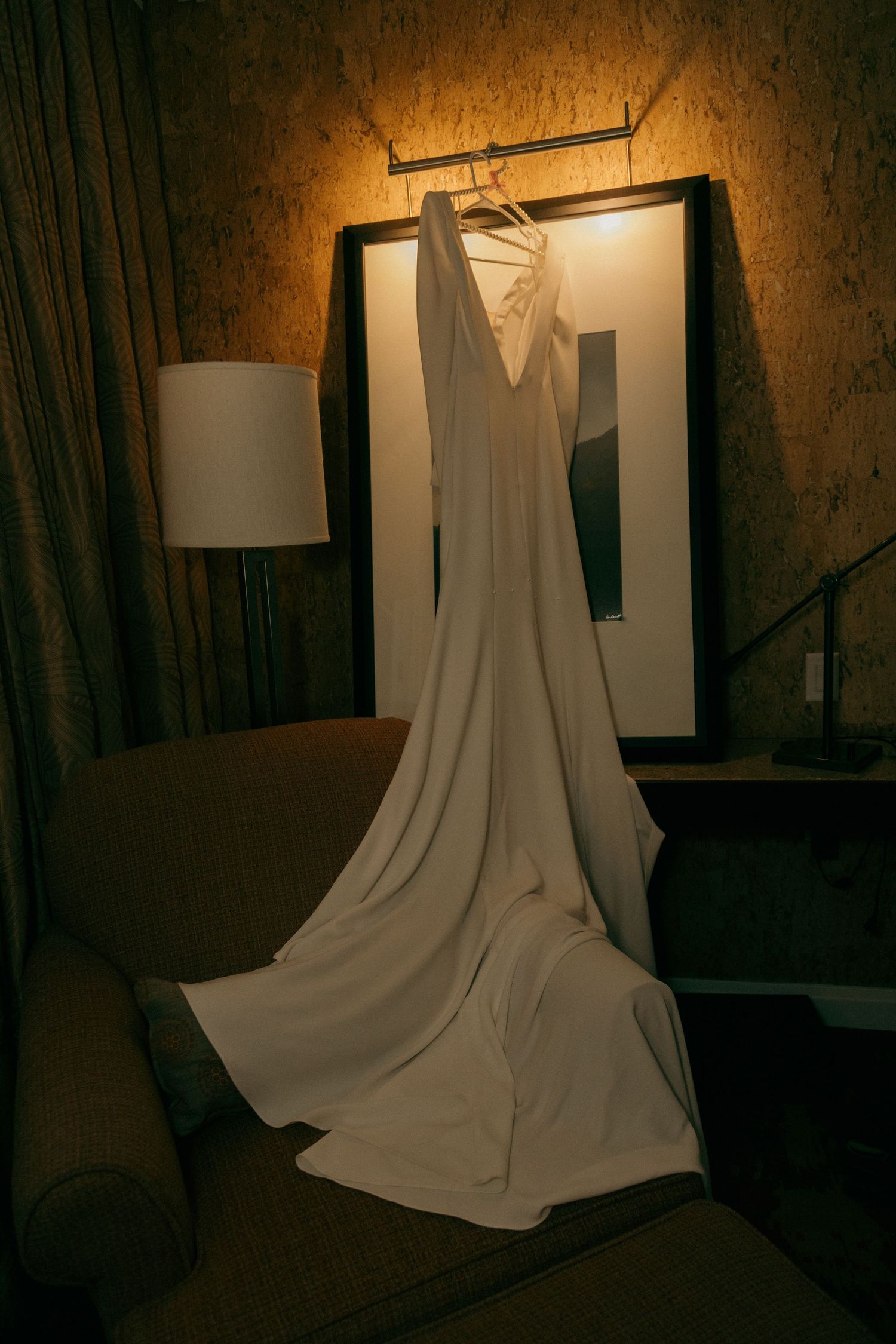 Elegant white wedding dress hanging on a hanger over a framed picture in a warmly lit hotel room.