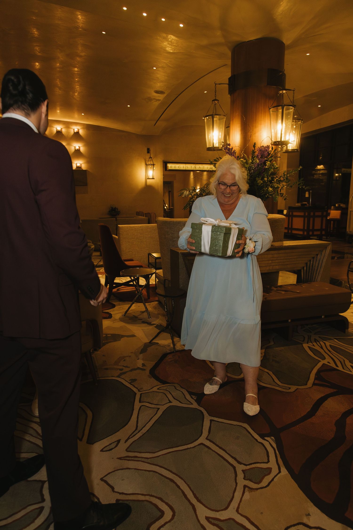 Elderly woman in light blue dress carrying a wrapped gift in an elegantly lit hotel lobby.