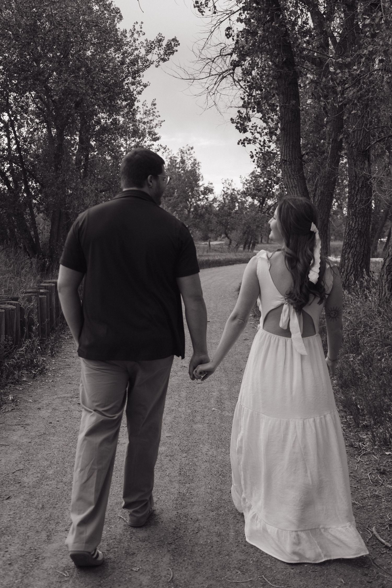Black and white photo of a couple holding hands walking down a tree-lined path, woman in white dress.