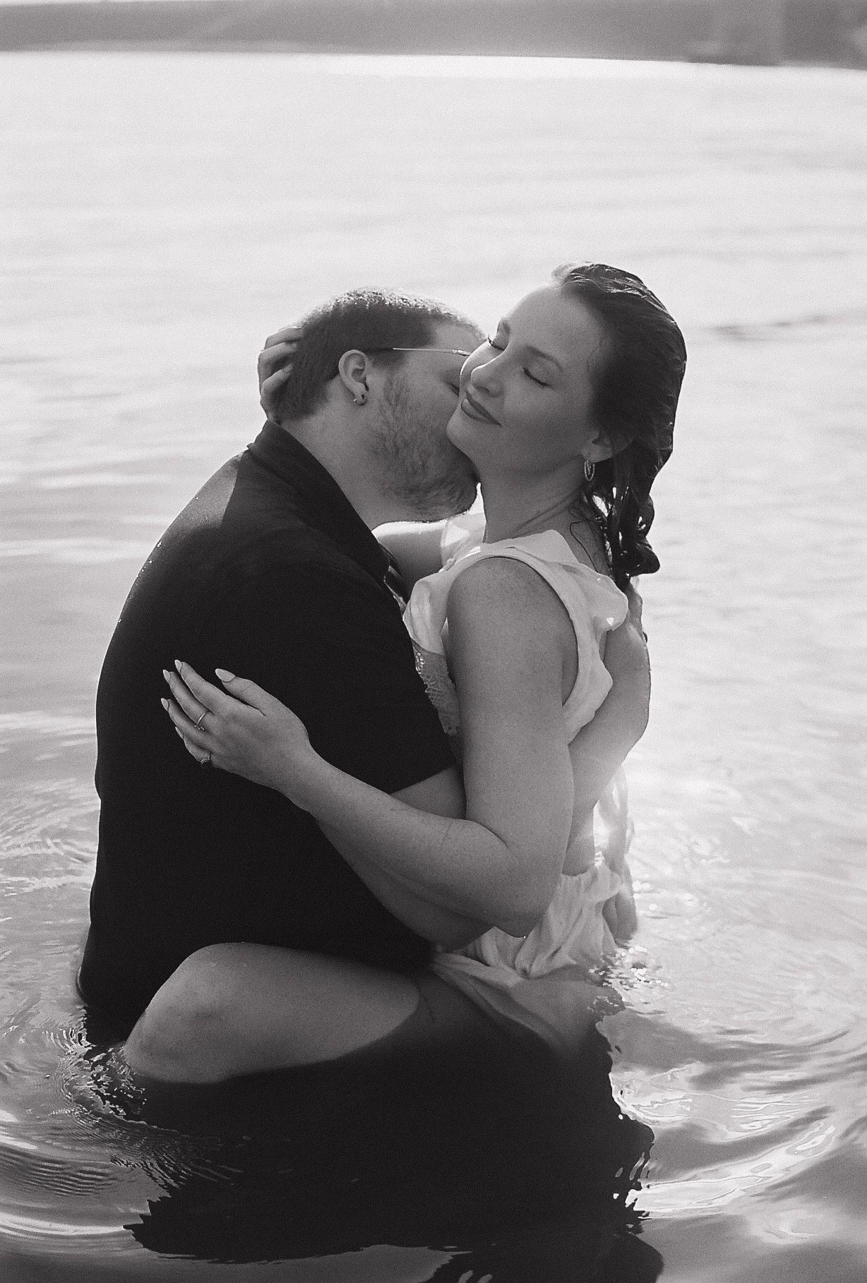 Black and white photo of couple sharing intimate embrace while standing in lake water together.