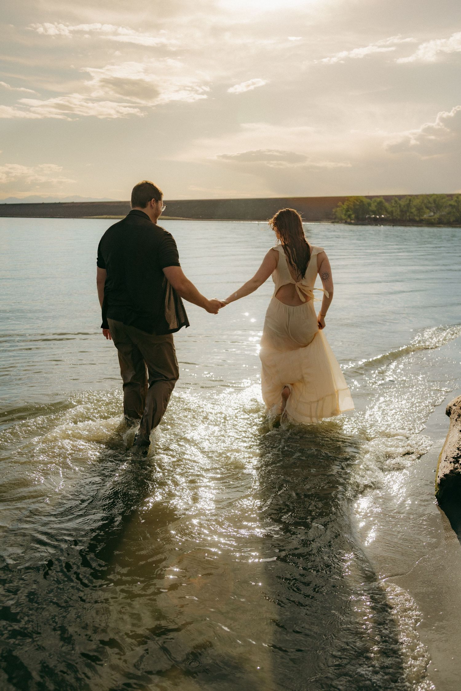 Couple walking hand in hand through gentle lake waves during a golden sunset.