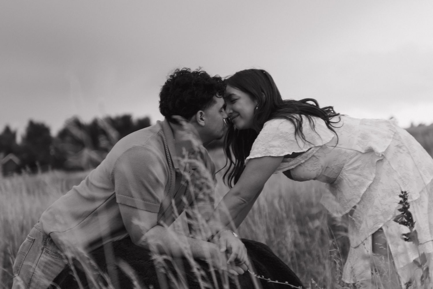 Black and white photo of couple sharing a tender almost-kiss among tall grass in field.