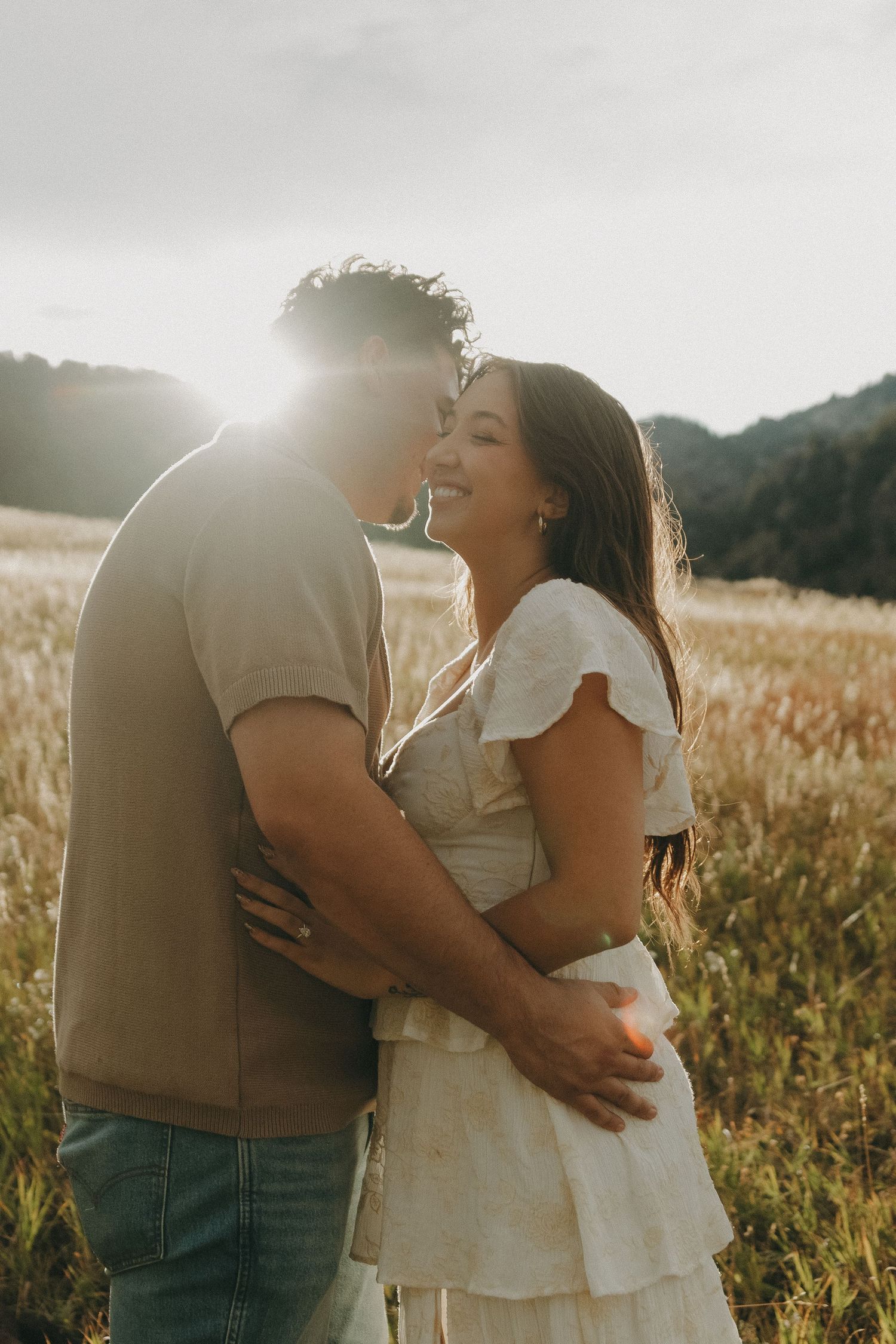 Couple embracing and laughing in sunlit golden meadow with mountains in background.