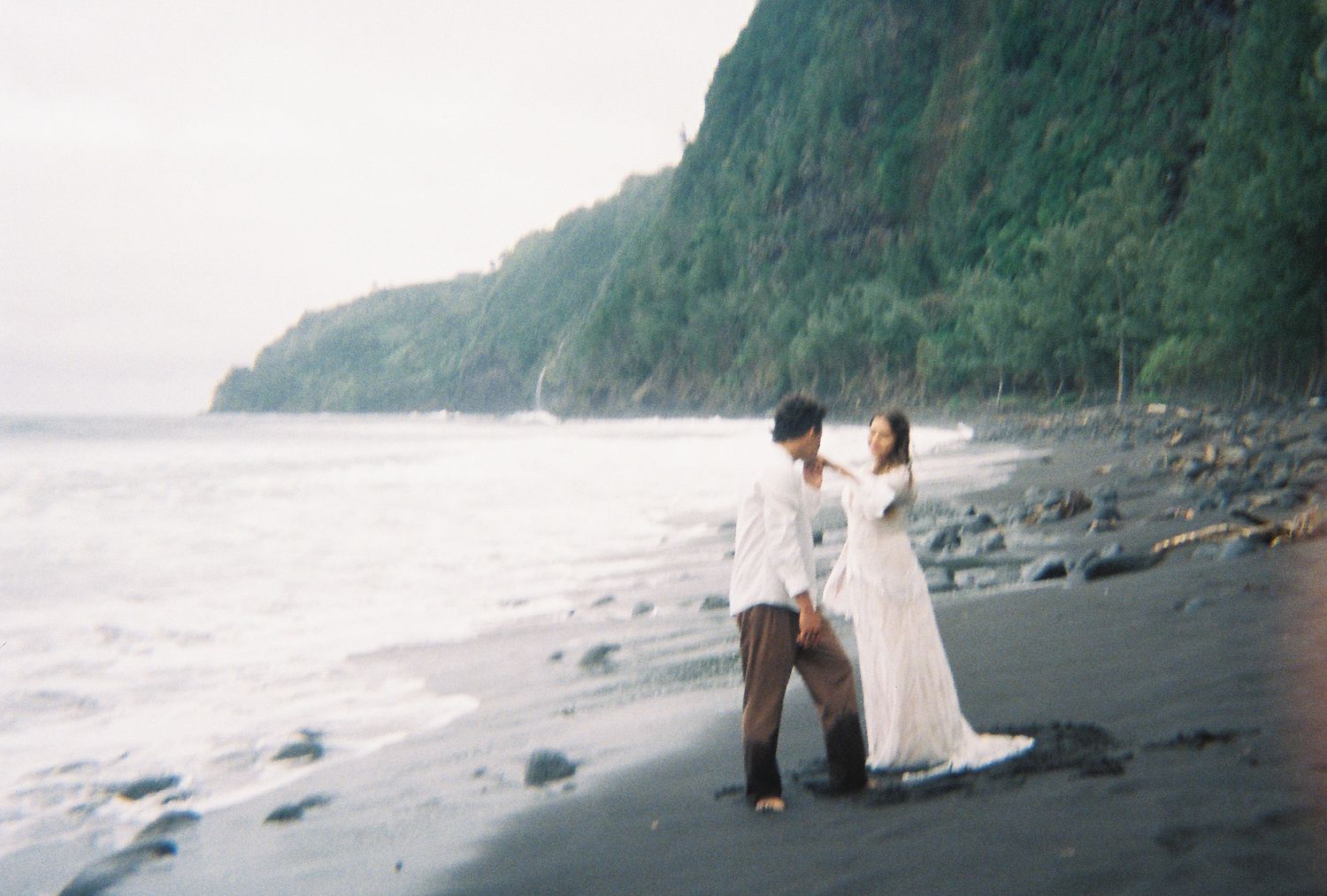 Couple in wedding attire dancing on a black sand beach with lush green cliffs and ocean waves in the background.