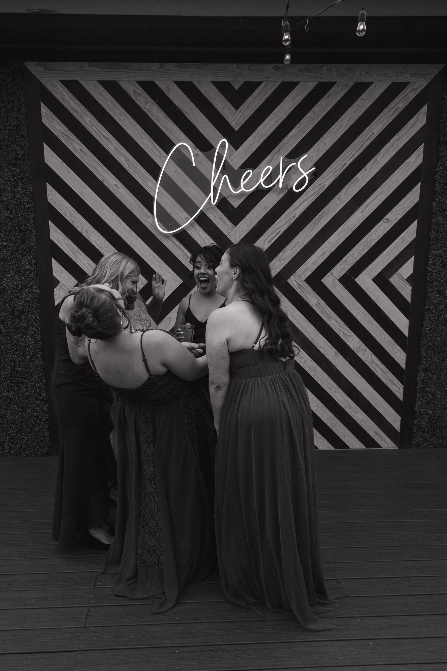 Bridesmaids in dark gowns laugh together in front of a Cheers neon sign backdrop.