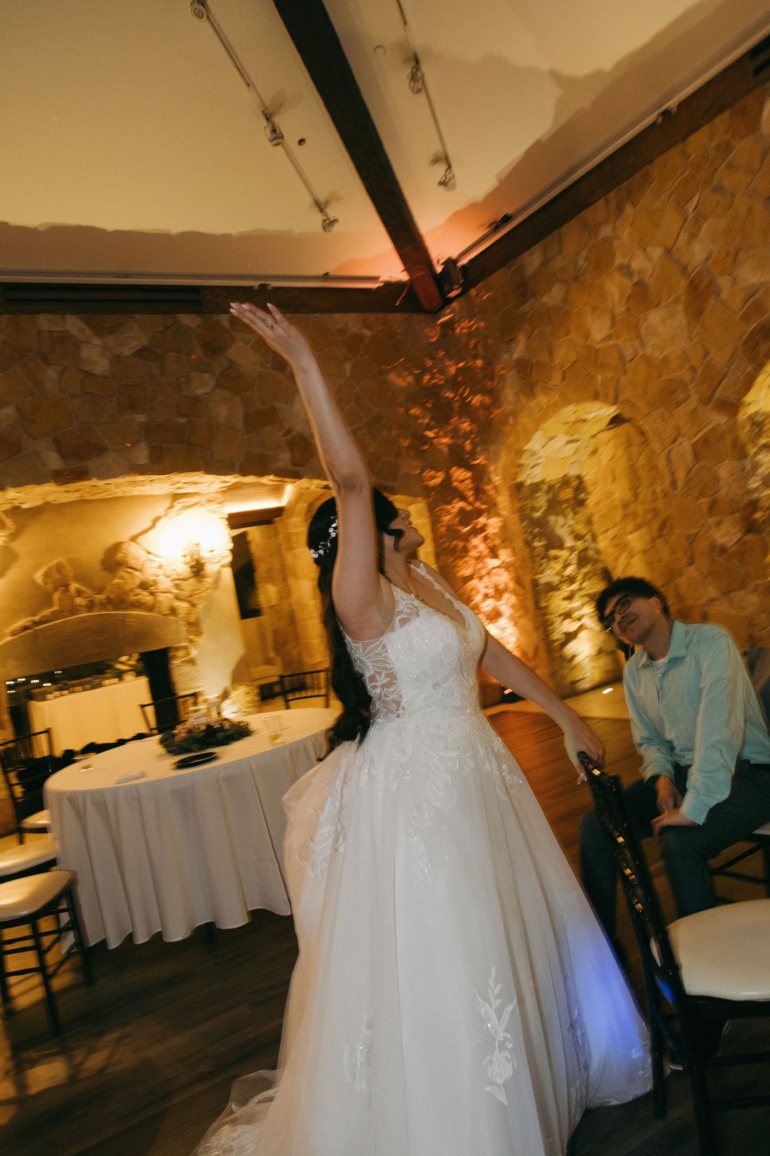 Bride raises arm joyfully in elegant lace gown inside rustic stone wedding reception venue.