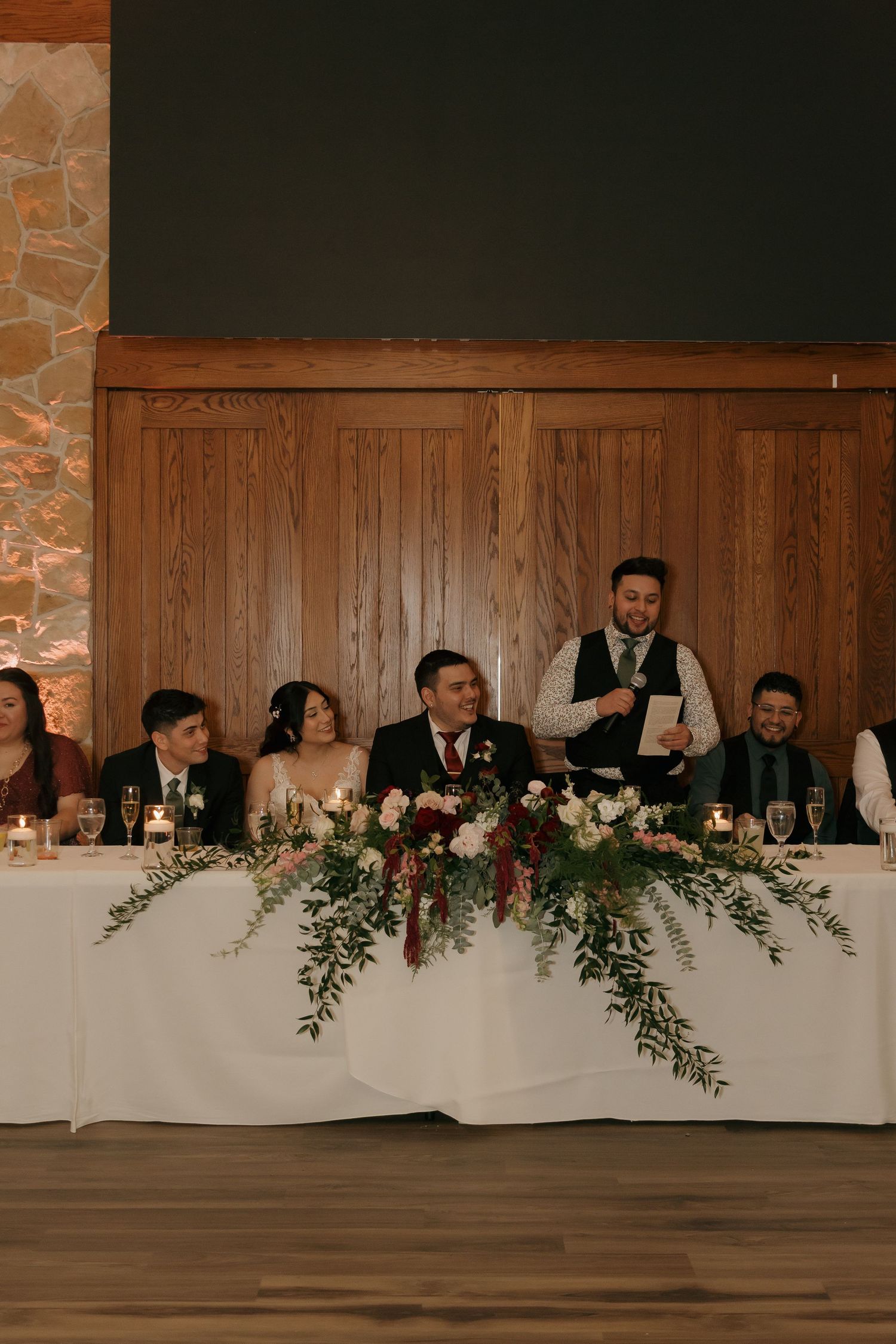 Wedding party seated at head table with lush floral garland during reception speech indoors.