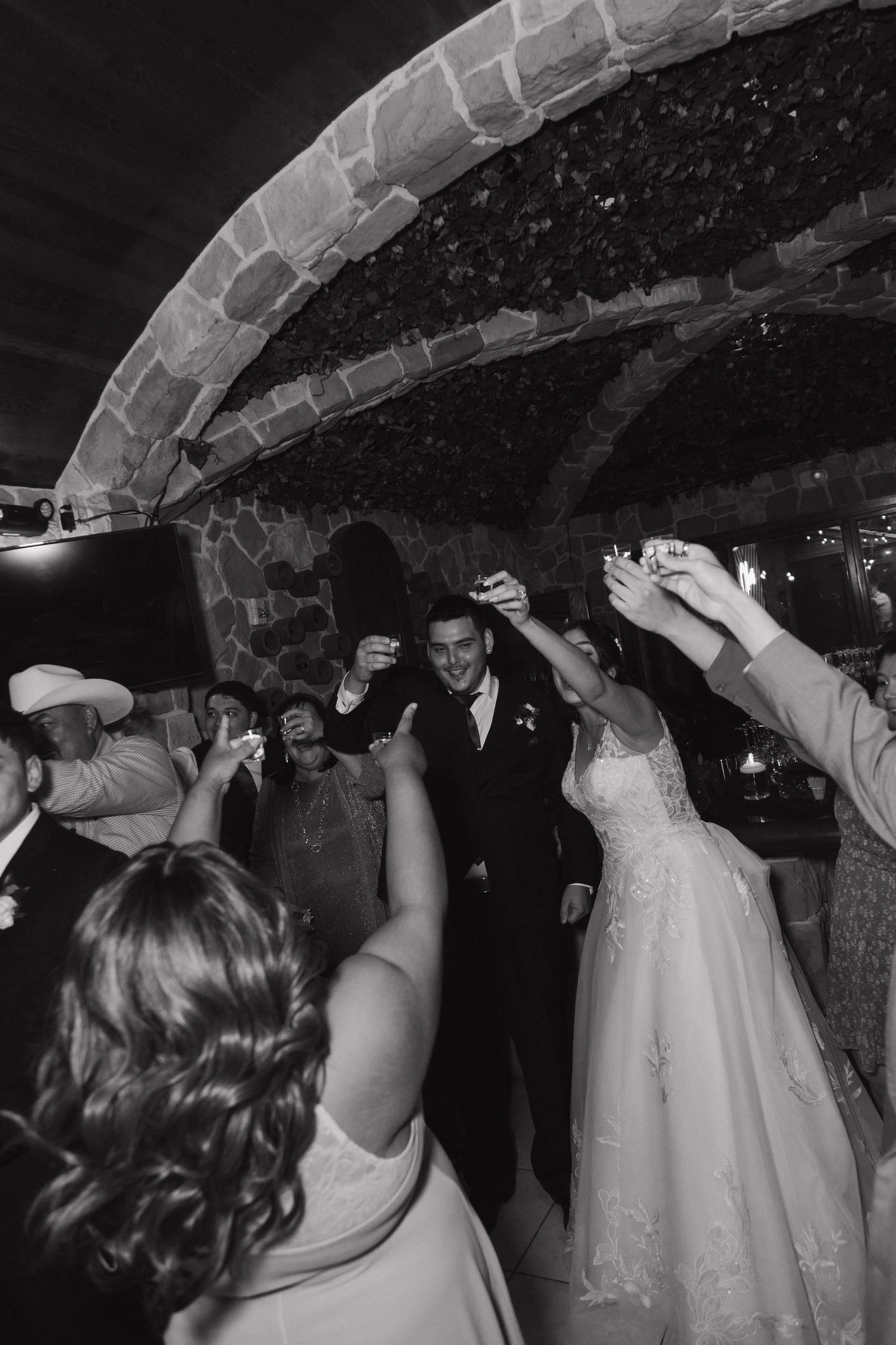 Black and white photo of bride and groom dancing surrounded by cheering guests under stone archway.