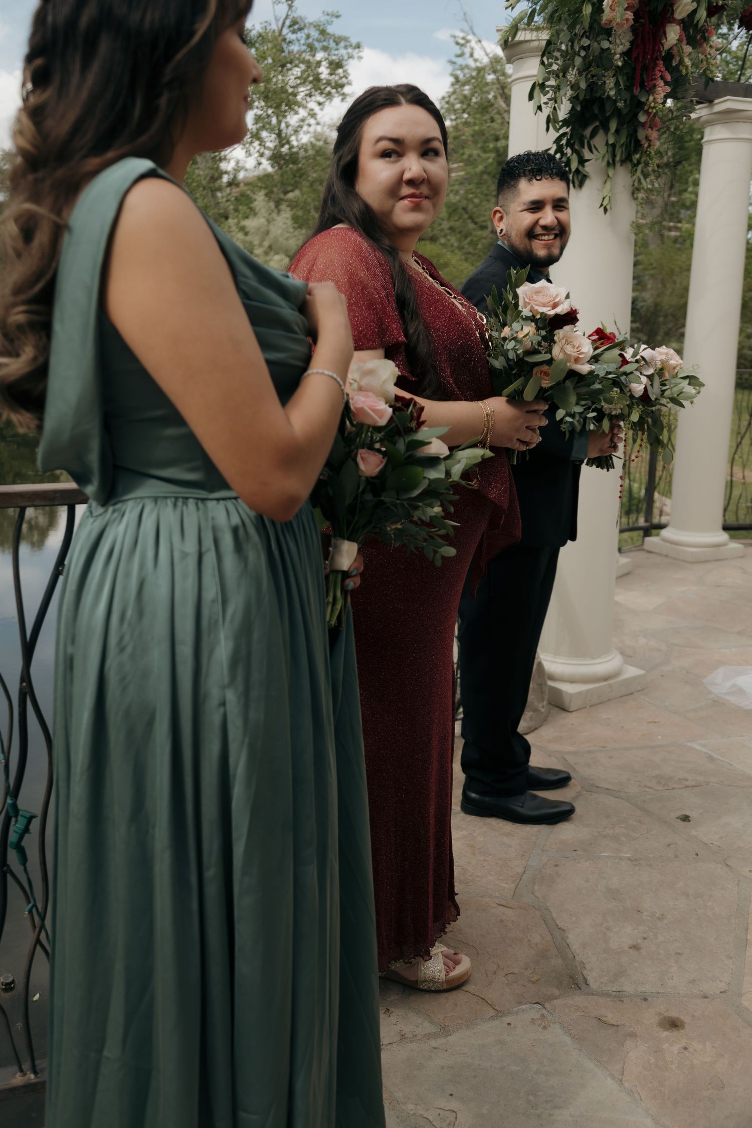 Smiling bridesmaid in burgundy dress holds bouquet beside groomsman at outdoor wedding ceremony with white columns.