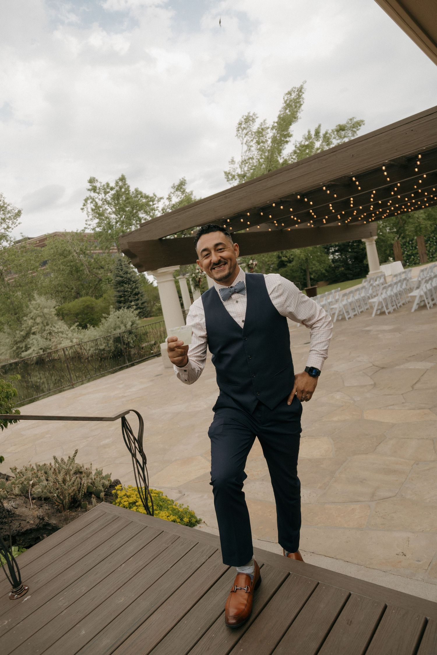 Smiling groom in navy vest and bow tie celebrates joyfully on a deck at an outdoor wedding venue.