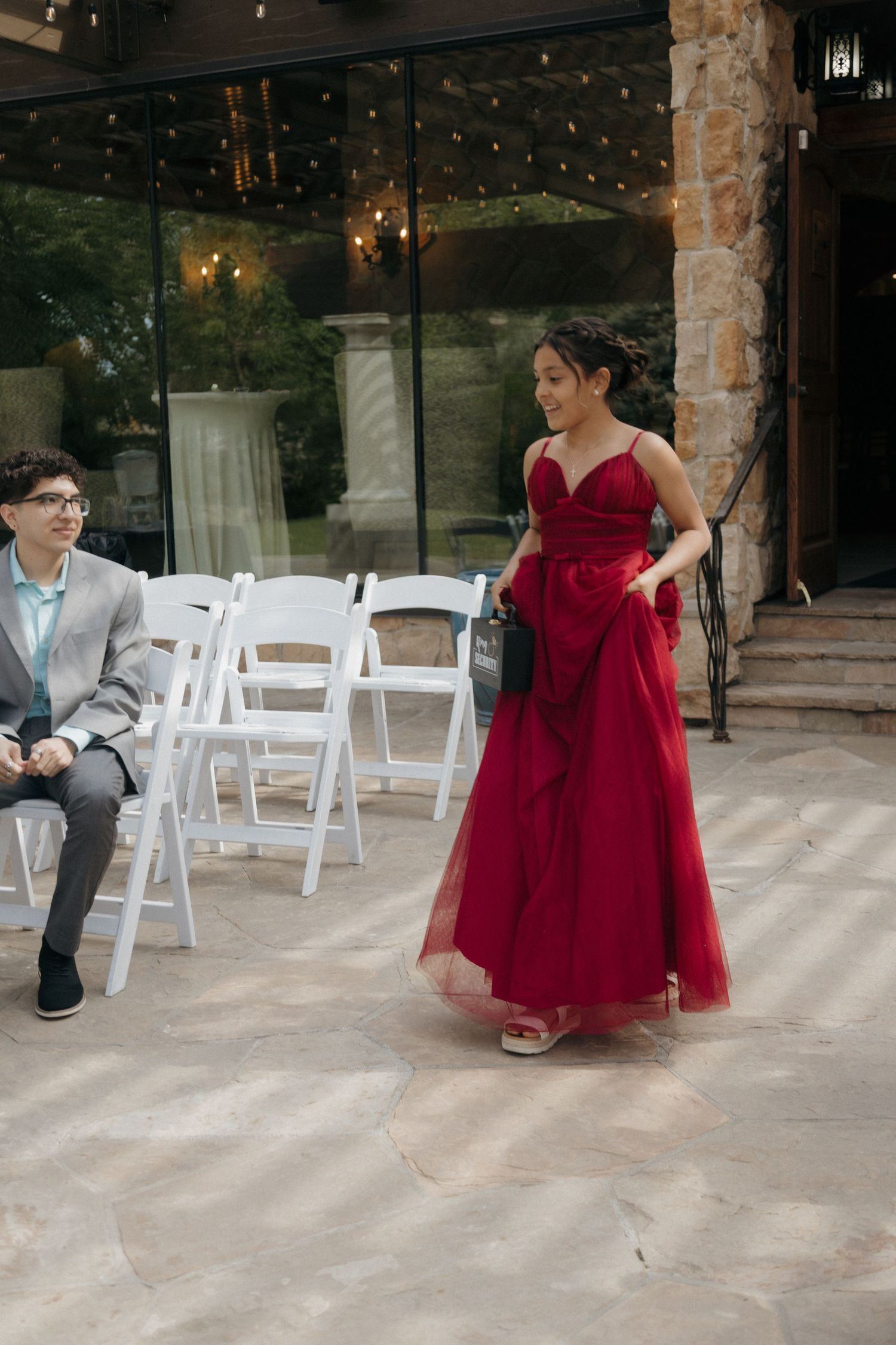 Bridesmaid in elegant red gown walks through outdoor wedding ceremony venue with white chairs.
