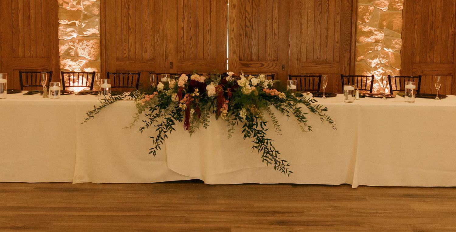 Elegant wedding head table with lush floral centerpiece of greenery and roses on white linen in a warm wood venue.