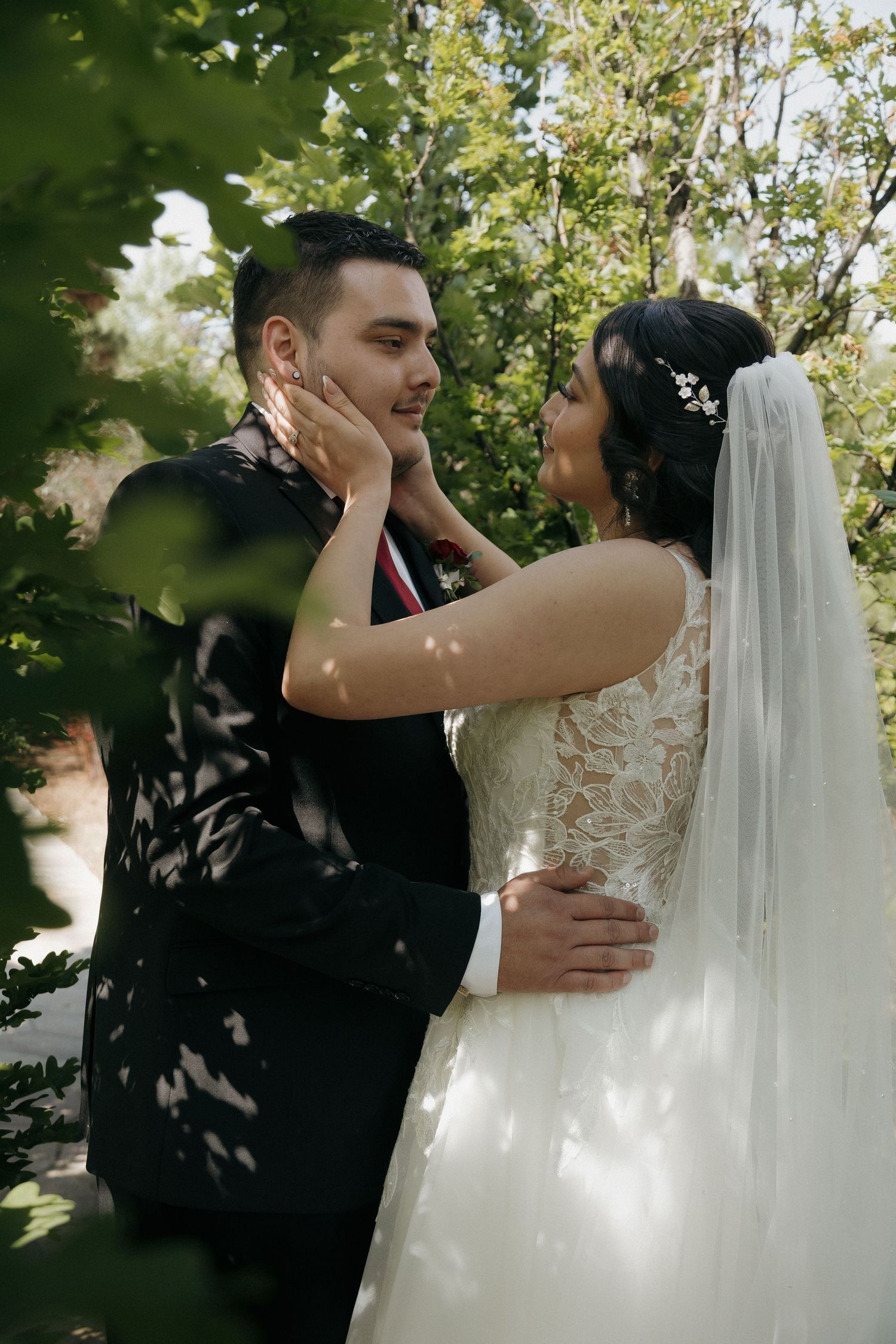 Bride tenderly holding grooms face as they embrace surrounded by lush green trees.