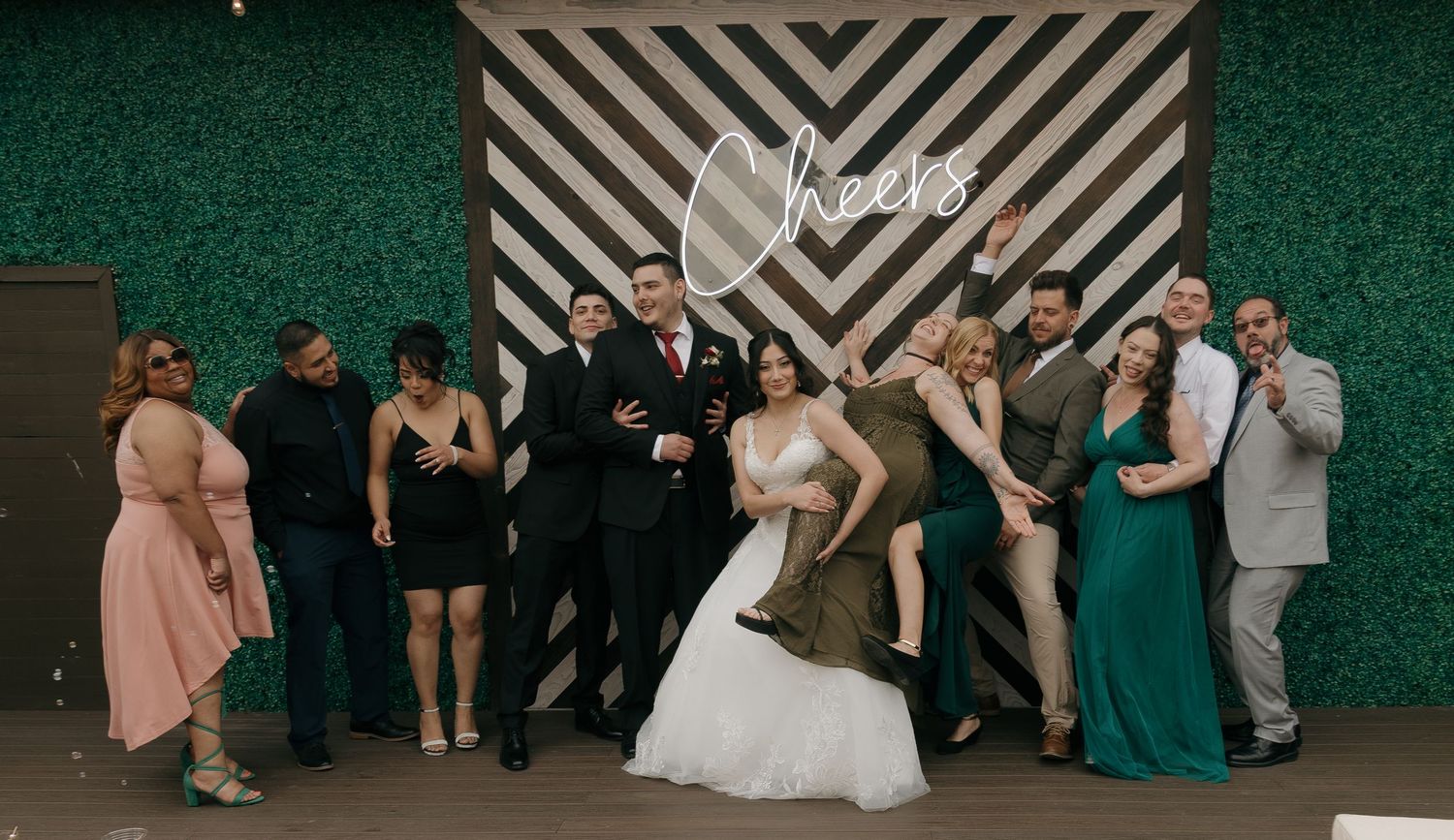 Wedding party group photo in front of striped backdrop with neon Cheers sign at festive reception celebration.