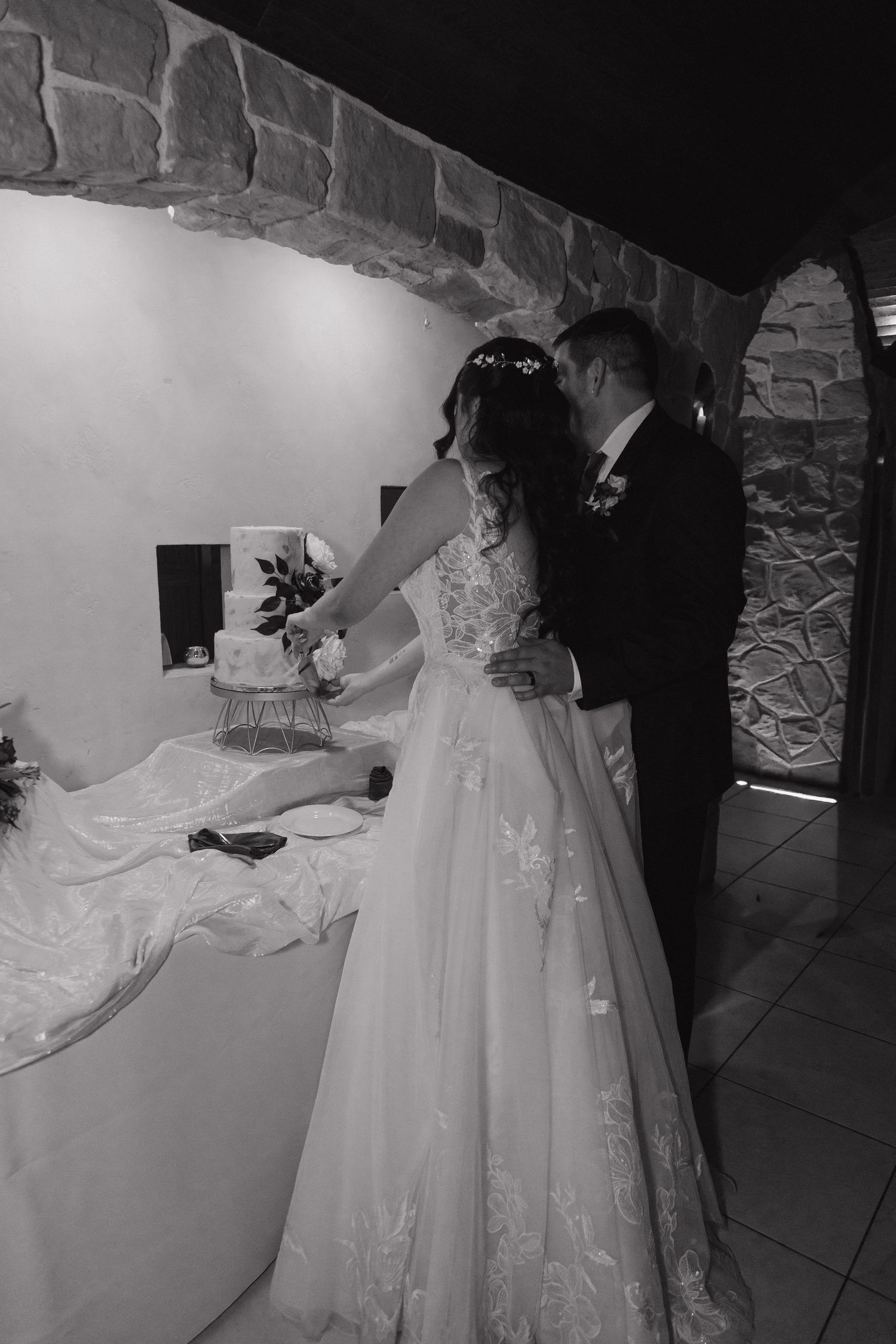 Bride and groom cut wedding cake together in elegant stone venue in black and white photo.