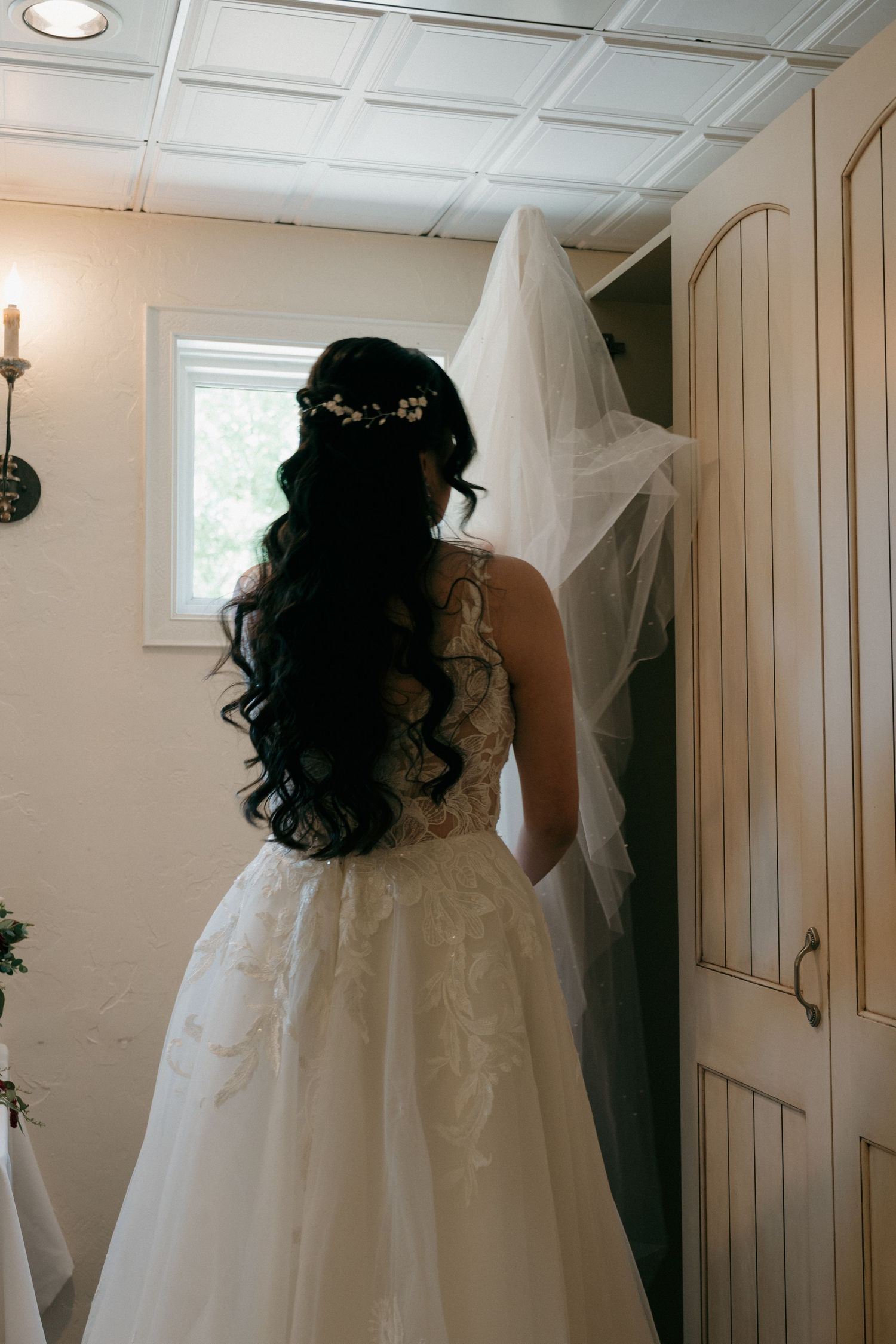 Bride in lace ball gown with long dark curly hair, floral crown, and veil viewed from behind.
