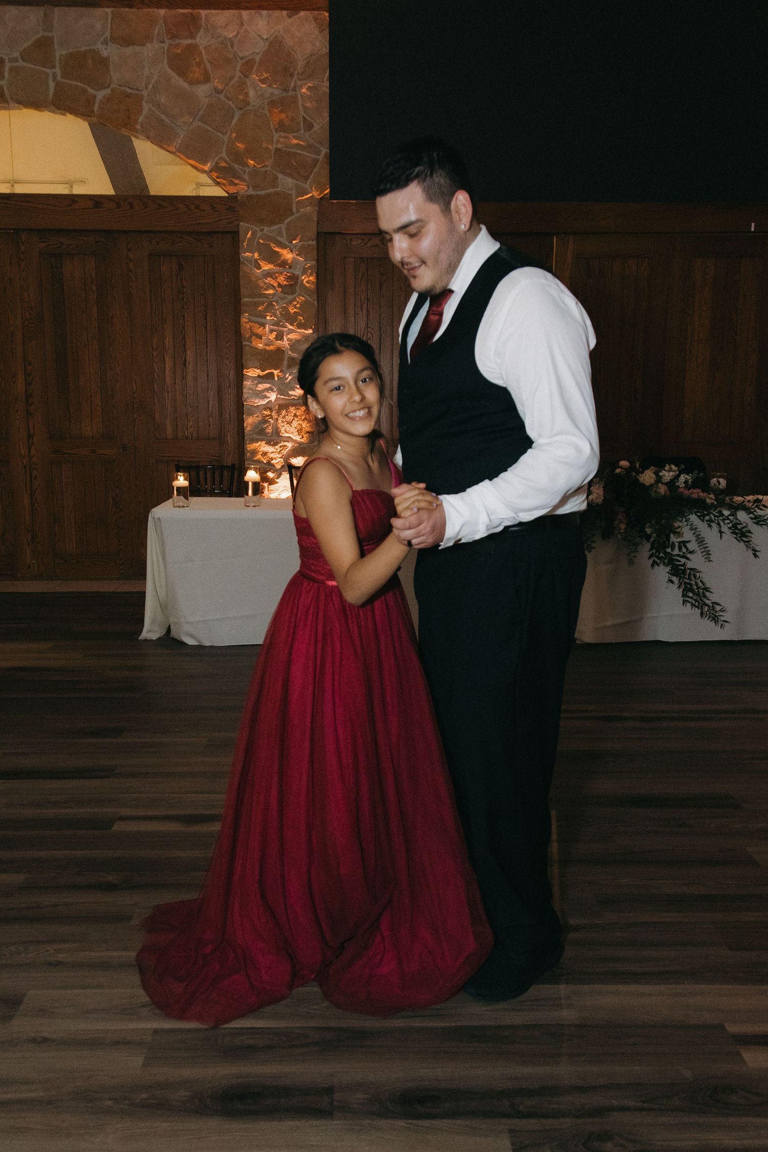 Groom in black vest dances with a young girl in a red ballgown at an elegant wedding reception.