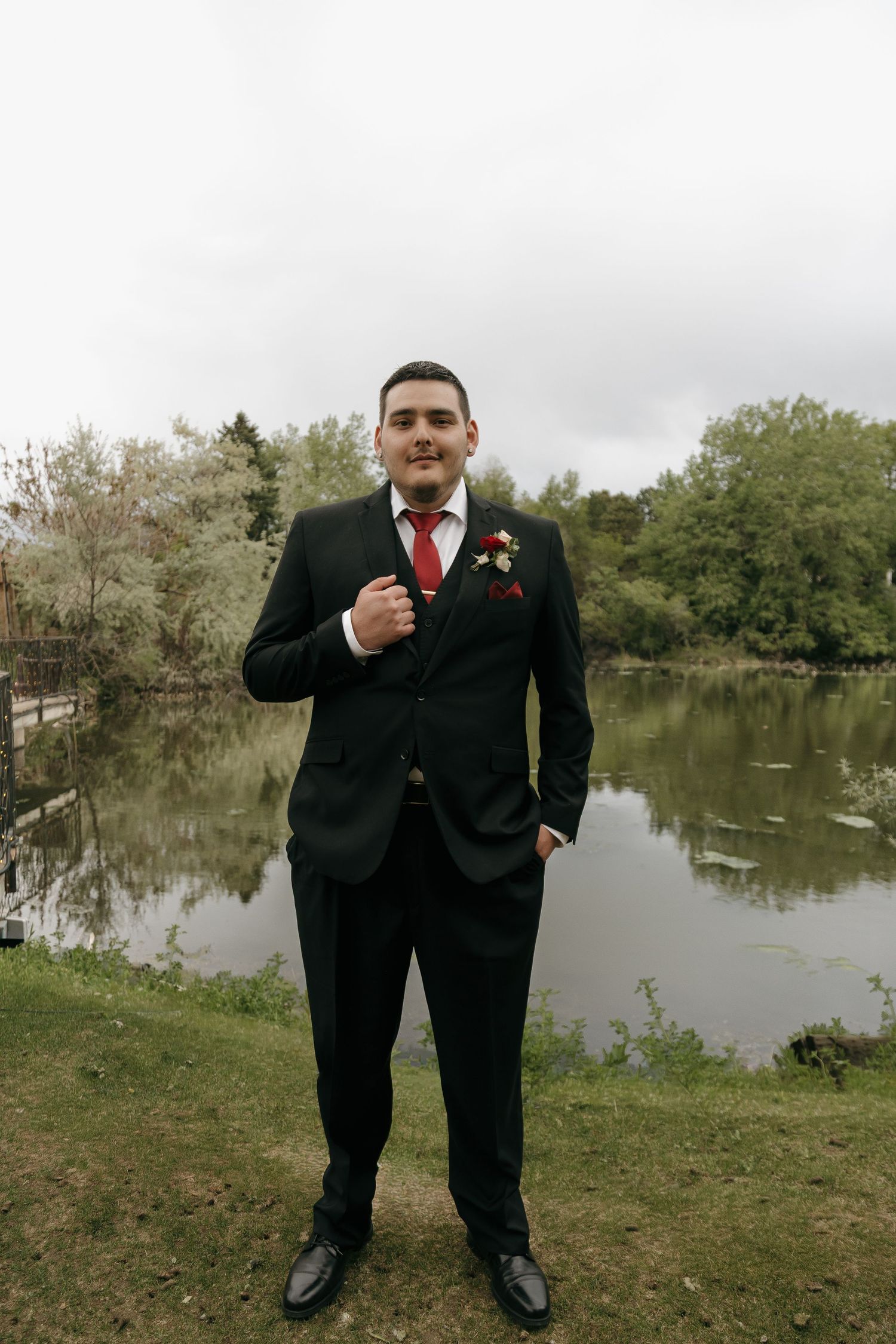 Dapper groom in black suit with red tie and boutonniere poses confidently by a serene pond.