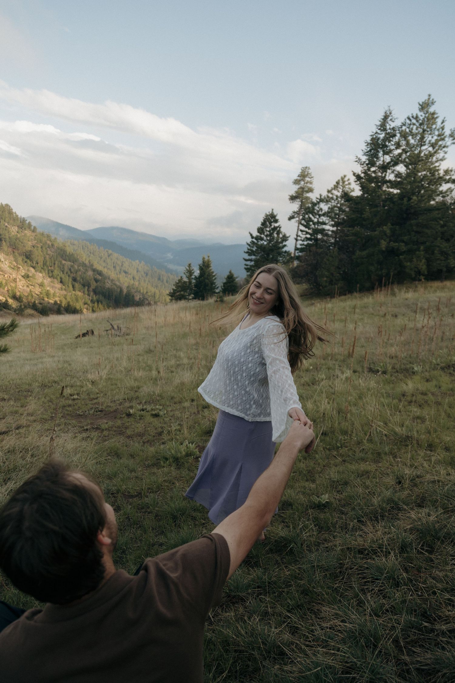 Woman laughing joyfully while holding hands with partner on a mountain meadow at dusk.