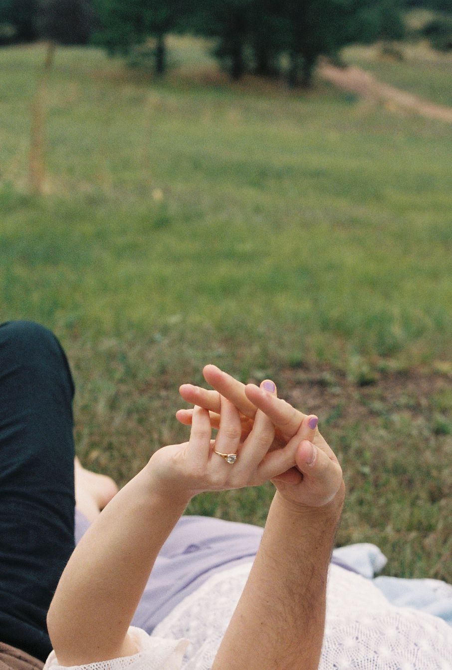 Two hands gently intertwined outdoors, one wearing a gold ring, with a lush green meadow in the background.
