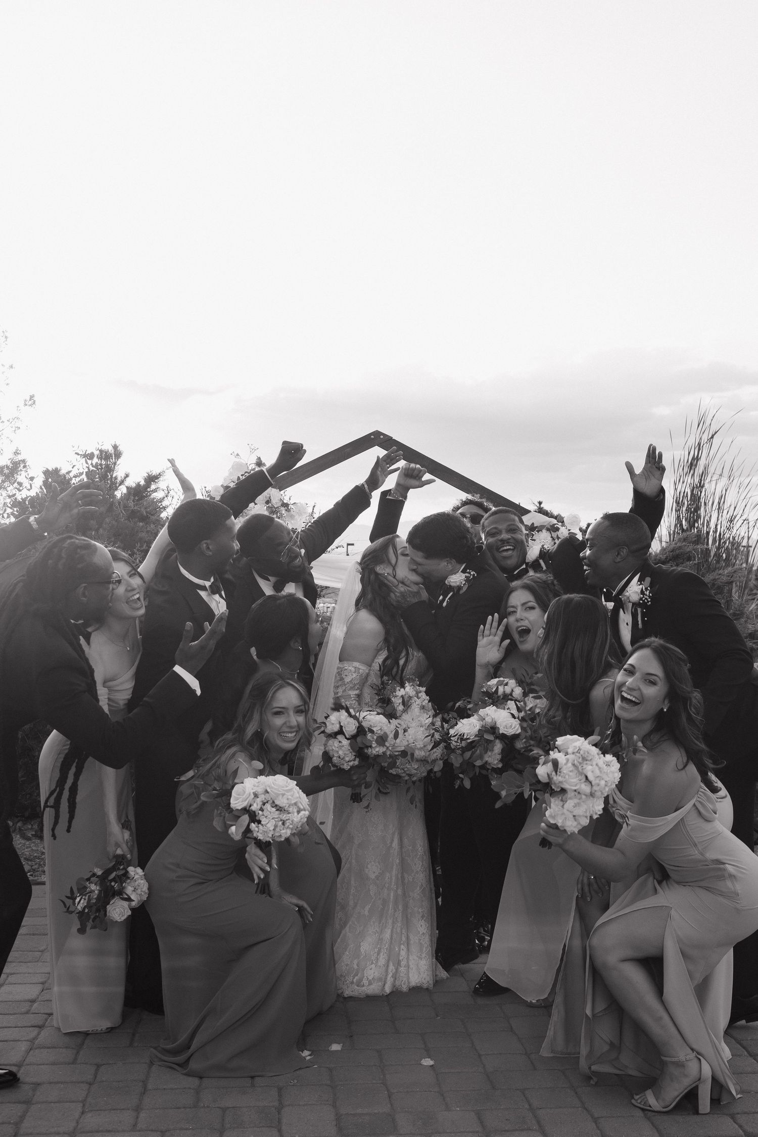 Joyful wedding party celebrates with bride and groom under arch in lively black and white photo.